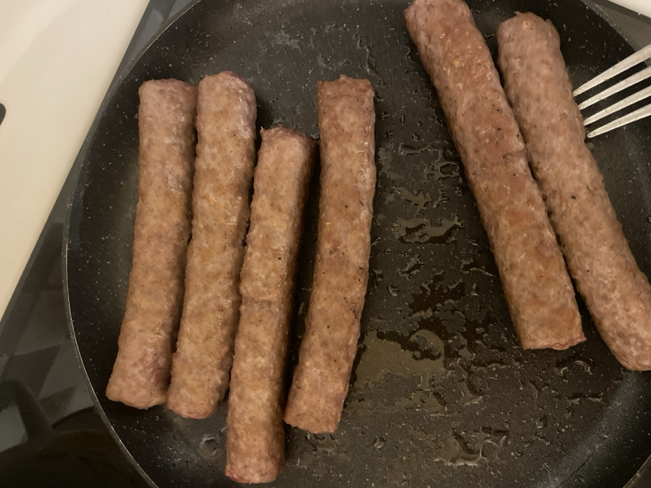 This image depicts a cooking scene in the city of Palež, Montenegro. Seven elongated, cylindrical meat items, consistent in appearance with sausages or ćevapi, are contained within a black, speckled non-stick frying pan. Five of these items are arranged horizontally in the upper-left portion of the pan, parallel to each other. The remaining two items are positioned diagonally in the lower-right section. The meat items exhibit a browned, grayish-pink exterior with visible searing marks. The pan's surface shows various droplets and streaks of cooking oil or grease. A silver-colored, four-tined fork is partially visible in the lower-right, its tines in contact with one of the diagonally positioned meat items. The frying pan rests on a dark stovetop, with a lighter, indistinct surface visible in the background.