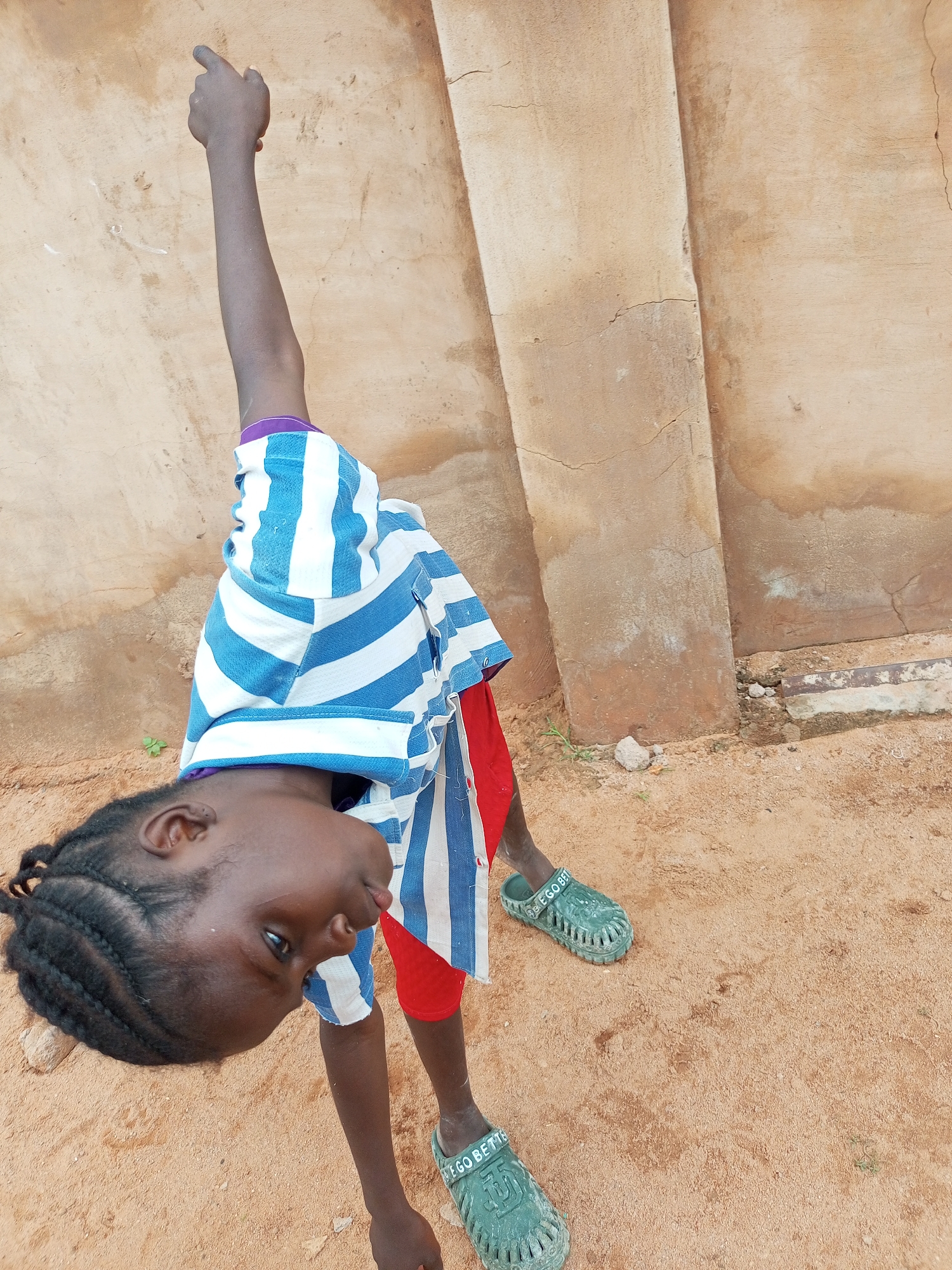A young child, possibly a girl, is positioned in a contorted pose, leaning downwards with their body at an angle. The child is wearing a blue and white striped t-shirt and red shorts, along with green clogs. One arm is extended upwards, with the index finger pointing. The child's face is turned downwards, looking towards the ground. The setting appears to be an outdoor environment with a sandy ground and a textured, light-colored wall in the background. The wall has visible cracks and a section with a metallic element embedded in the concrete base. The lighting suggests it is daytime. The image appears to have been taken in Tempure, Nigeria.