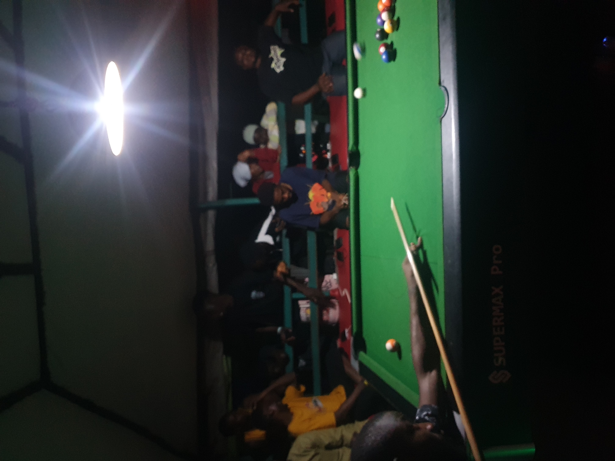 This media file depicts a group of people playing pool in what appears to be an indoor setting, possibly a bar or a recreational area, at night. The lighting is dim, with a bright, circular light fixture casting a strong glare on the left side of the frame. The green pool table dominates the right side of the image, with several colored pool balls visible, including what appears to be a white cue ball in motion.
Several individuals are gathered around the pool table, some actively engaged in the game, while others seem to be observing. One person, wearing a dark t-shirt with a graphic print, is leaning over the table, appearing to be taking a shot with a pool cue. Another individual in a blue t-shirt with an orange design is seated nearby, looking towards the table. In the background, other figures are visible, some standing and others seated, in various states of engagement. The overall atmosphere appears casual and social.
The image is oriented vertically, with the pool table running along the right edge. The scene is dark, suggesting it is nighttime. There are no clear indications of weather conditions. The presence of a pool table and the social gathering suggests a recreational or entertainment venue.
Visible text includes "SUPERMAX Pro" printed vertically on the dark side of the pool table. There are no other discernible text elements in the image. The emotional tone is difficult to ascertain due to the low quality and lighting, but the activity suggests a relaxed and possibly competitive social gathering. The location context of Lokoja, Nigeria, suggests this could be a local establishment.