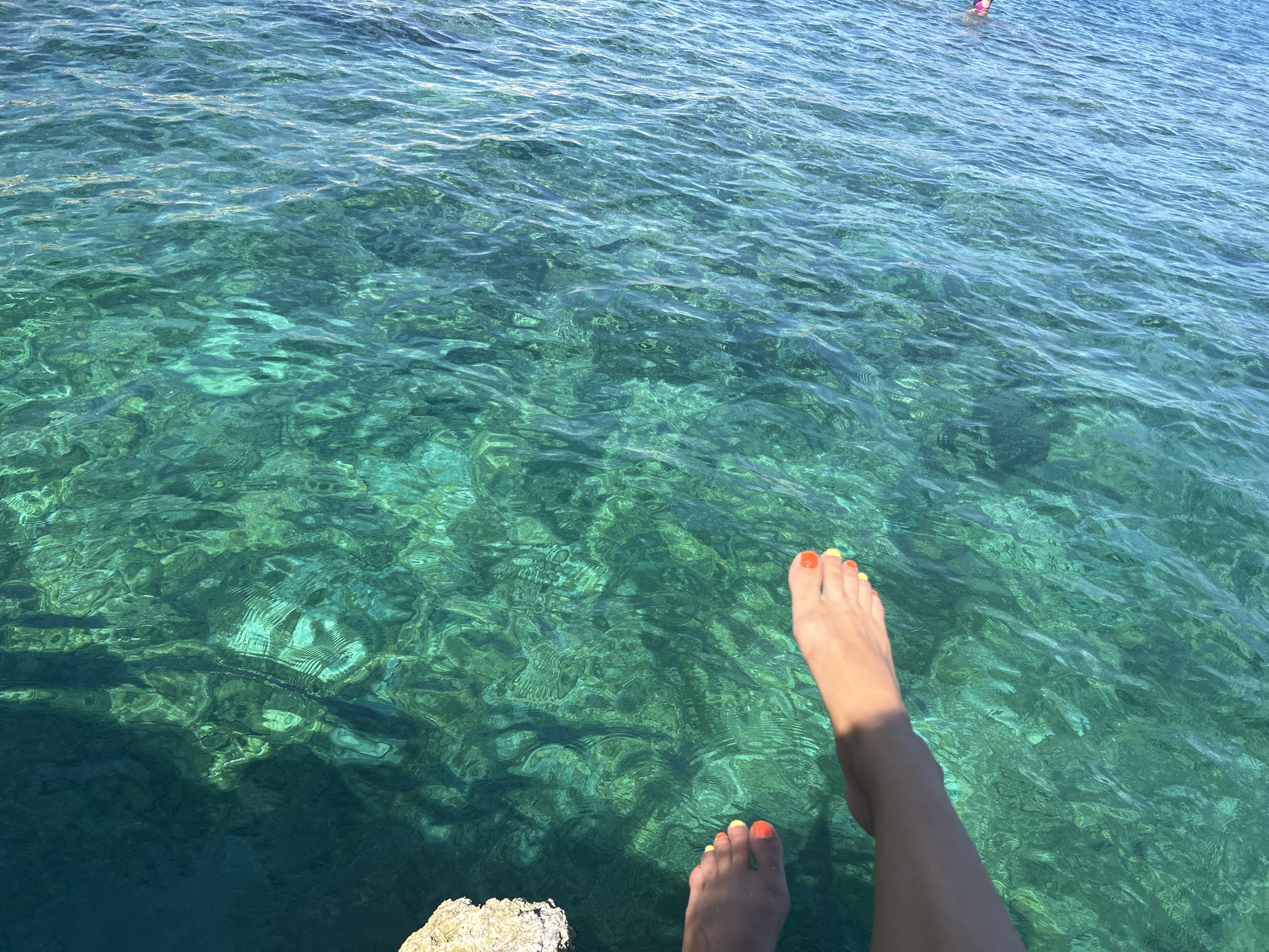 In the crystal-clear turquoise waters of Cala Lliteres, Spain, a person's feet, adorned with bright orange and yellow nail polish, dangle above the surface. The sun-drenched water reveals the textured seabed below, creating dappled patterns of light and shadow. In the distance, another individual is partially visible, swimming or floating in the refreshing sea. The scene evokes a sense of leisure and the serene beauty of a Mediterranean summer.