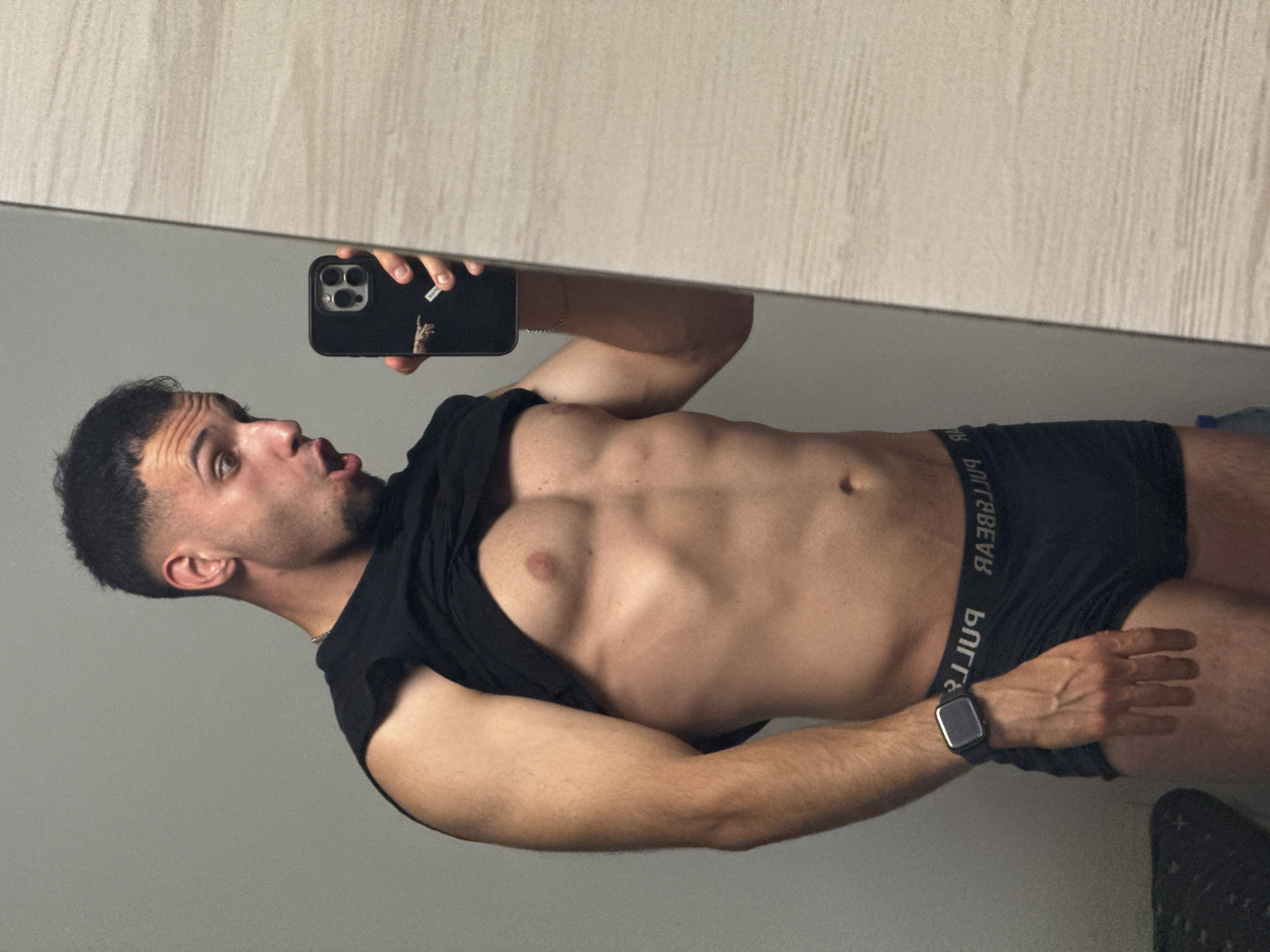 A youthful male, likely in his late teens or early twenties, is depicted taking a mirror selfie indoors in Shumen, Bulgaria. The image shows him horizontally, reflected in a large, clear mirror. He has short, dark hair and light facial stubble. He is dressed in a black t-shirt, which is pulled up to reveal his muscular torso and visible abdominal muscles. Below his shirt, he wears black boxer briefs with the brand "PULL&BEAR" visible in reverse on the waistband. A dark-colored smartwatch is on his left wrist, and a subtle silver chain adorns his neck.
He holds a black smartphone, featuring multiple camera lenses, in his right hand, angled to capture the selfie. His expression is animated, with his mouth forming an open "O" shape, conveying surprise or playfulness. The upper portion of the mirror's reflection shows a light-colored wooden or laminate surface, suggesting furniture or wall paneling. The area behind the camera, reflected in the lower part of the mirror, appears to be a plain light grey wall. A small white sticker on his phone case, visible in the reflection, shows a peace sign or "V" gesture made with fingers. The lighting is even, indicating an indoor setting.