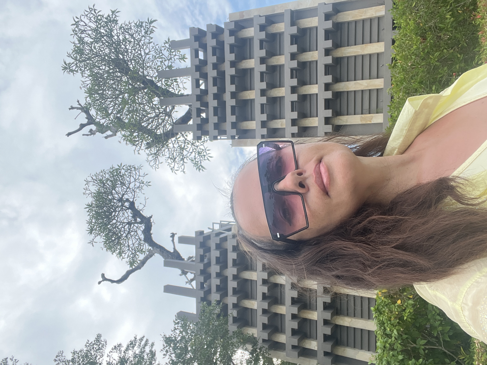 A low-angle, medium shot captures a woman in sunglasses posing for a selfie with an unusual architectural structure and trees in the background. The woman is positioned in the foreground, with her face and upper body filling the lower portion of the frame. She has long, dark, wavy hair and is wearing oversized, square-shaped sunglasses with a gradient pink tint. Her lips are slightly parted, and she appears to be looking directly at the camera. She is wearing a light yellow, collared shirt.
Behind her, a striking gray, multi-tiered structure with a geometric design resembling stacked blocks forms the central focus of the background. Two distinct trees, sculpted and appearing almost artificial, emerge from the tops of the structures. Their branches are bare in some areas, with sparse green foliage in others. The sky above is cloudy with patches of blue. In the lower right and left edges of the frame, lush green bushes are visible. The overall scene suggests an outdoor location, possibly a park or a unique garden area within Kuta Selatan, Indonesia, given the distinctive architectural style and tropical-looking trees.