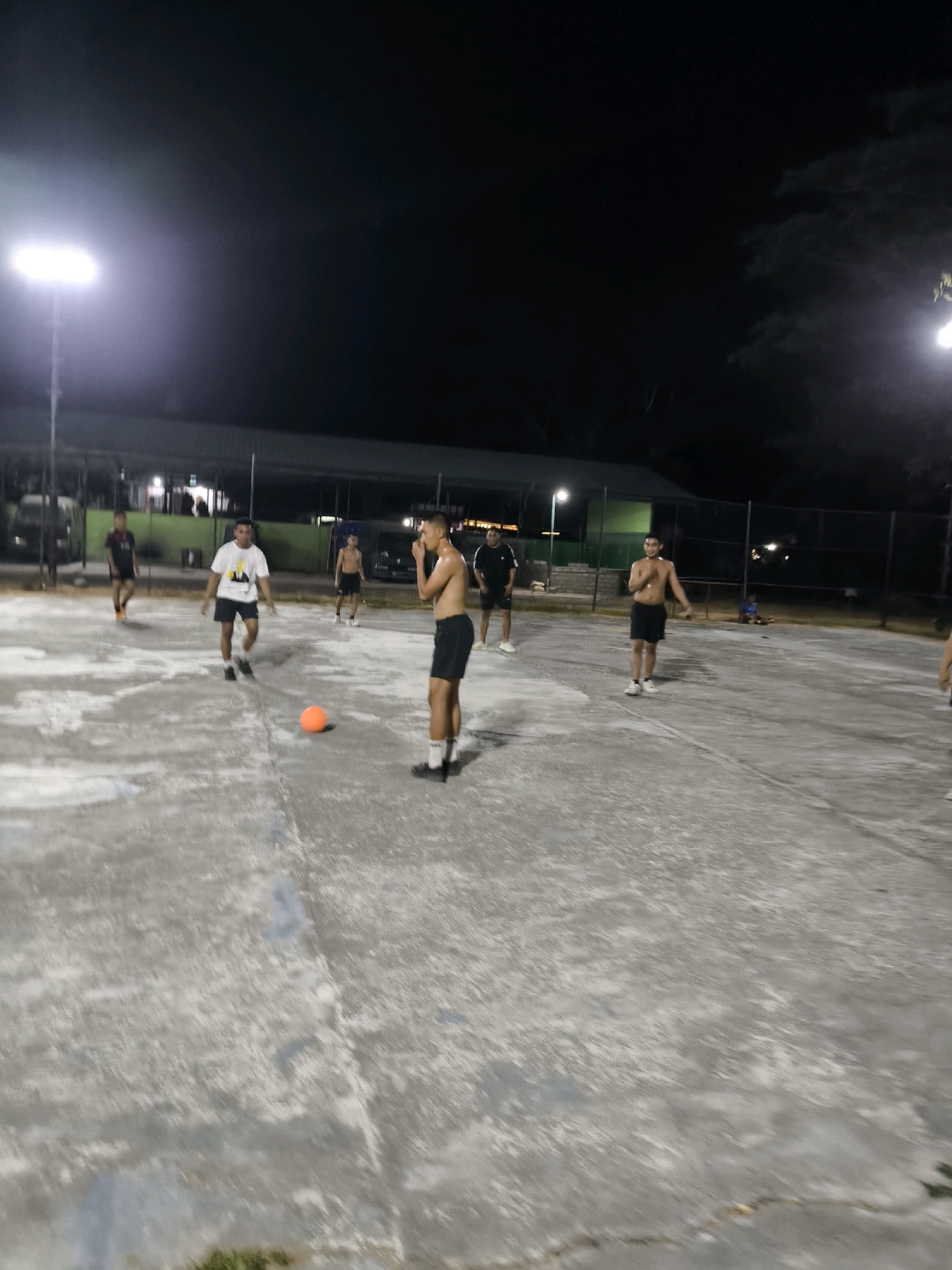 The image depicts an outdoor sports court at night, illuminated by floodlights. Several young men are present on the court, some actively engaged in a game, while others appear to be observing or resting. One individual in the foreground, shirtless and wearing black shorts, is looking towards the right of the frame with his hands near his face, possibly signaling or in a defensive stance. Another young man in a white t-shirt and black shorts is walking towards an orange ball on the ground. In the background, other individuals are visible, some playing a sport, and others standing or sitting. The court surface appears to be concrete, with some grass visible at the edges. In the far background, there are structures that suggest a community sports facility, possibly within an urban or suburban setting. The overall atmosphere is casual and recreational.