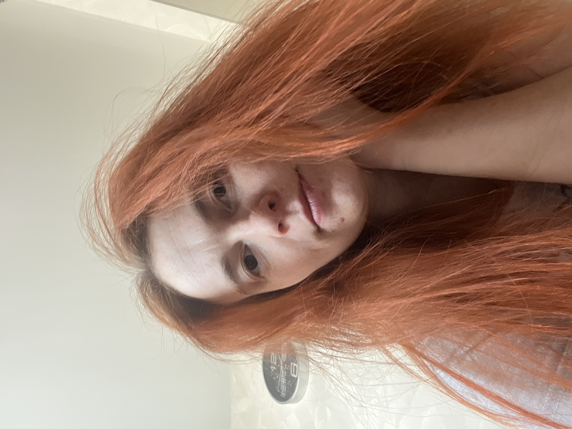 A close-up, rotated selfie shows a person with reddish-orange, medium-length hair. The individual has fair skin and their face is the focal point. In the background, slightly out of focus, there is a wall clock and the wall is white. The location is unknown.