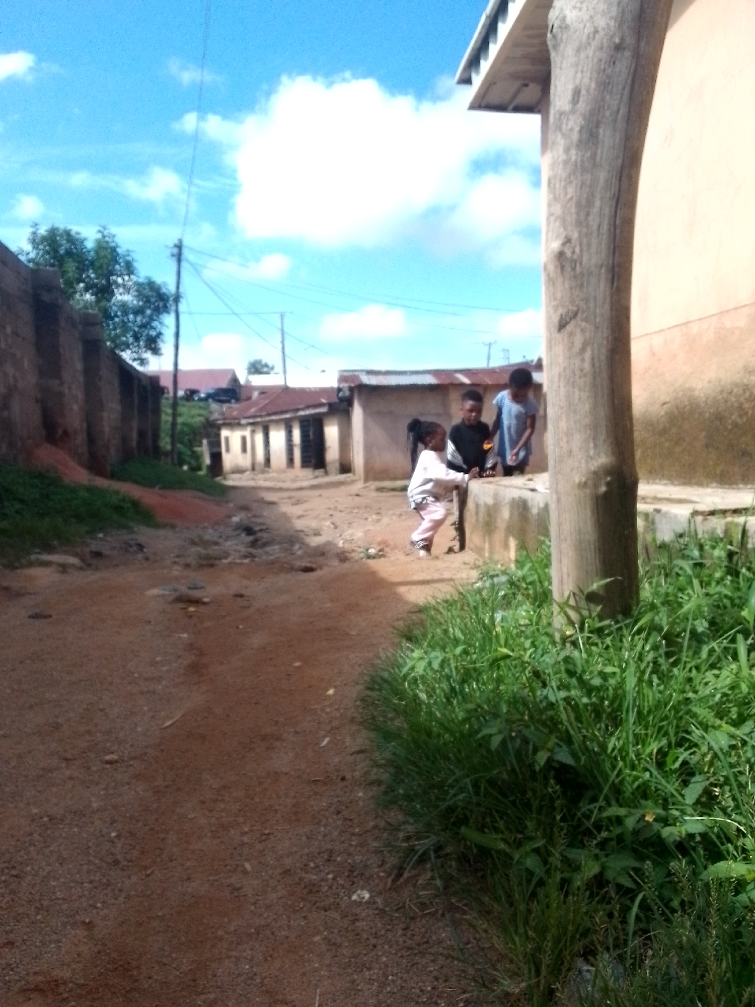 In an outdoor, unpaved street scene, three children are present. A young girl with dark hair pulled back in a ponytail is in the foreground, wearing a light-colored long-sleeved shirt and pink pants, and appears to be walking. Behind her, two slightly older children are standing near a low, light-colored wall. One child, wearing a black shirt with a white design, looks towards the girl. The other child, wearing a blue and white striped shirt, is partially obscured. The environment is characterized by a dusty, reddish-brown dirt road. To the left, a rustic brick wall runs along the side of the road, with green vegetation growing at its base. In the background, several buildings with reddish-brown roofs are visible, along with utility poles and power lines against a bright blue sky with scattered white clouds. A large, rough-textured wooden post stands prominently to the right of the frame. Lush green grass and plants are abundant in the foreground to the right.