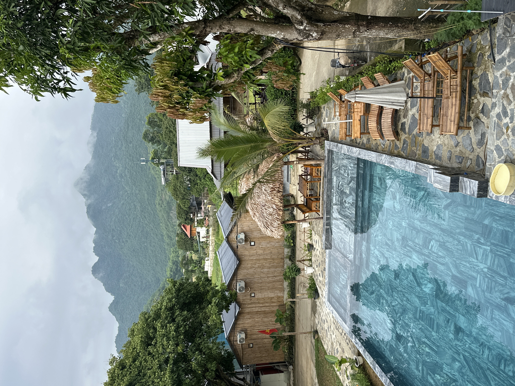 The image captures a serene resort scene in Xóm Khán, Vietnam, under an overcast sky. A clear, blue swimming pool with a marbled tile bottom dominates the foreground, reflecting the surrounding greenery and structures. Several empty wooden lounge chairs with grey cushions are neatly arranged around the pool. To the left of the pool are three identical bamboo huts with pitched, dark grey roofs and visible air conditioning units. A Vietnamese flag flies on a flagpole in front of these huts.
Behind the huts, a pathway leads to a cluster of buildings with metal roofs. A thatched-roof pavilion with wooden tables and chairs sits between the huts and a palm tree, offering a shaded seating area. Lush green trees and foliage surround the resort, with a large tree on the right side of the frame providing shade. In the background, a majestic mountain range, shrouded in mist, rises above the trees and buildings, indicative of the region's natural beauty. A motorbike is parked near the large tree on the right. The overall impression is one of a tranquil retreat, nestled within a picturesque natural landscape.
