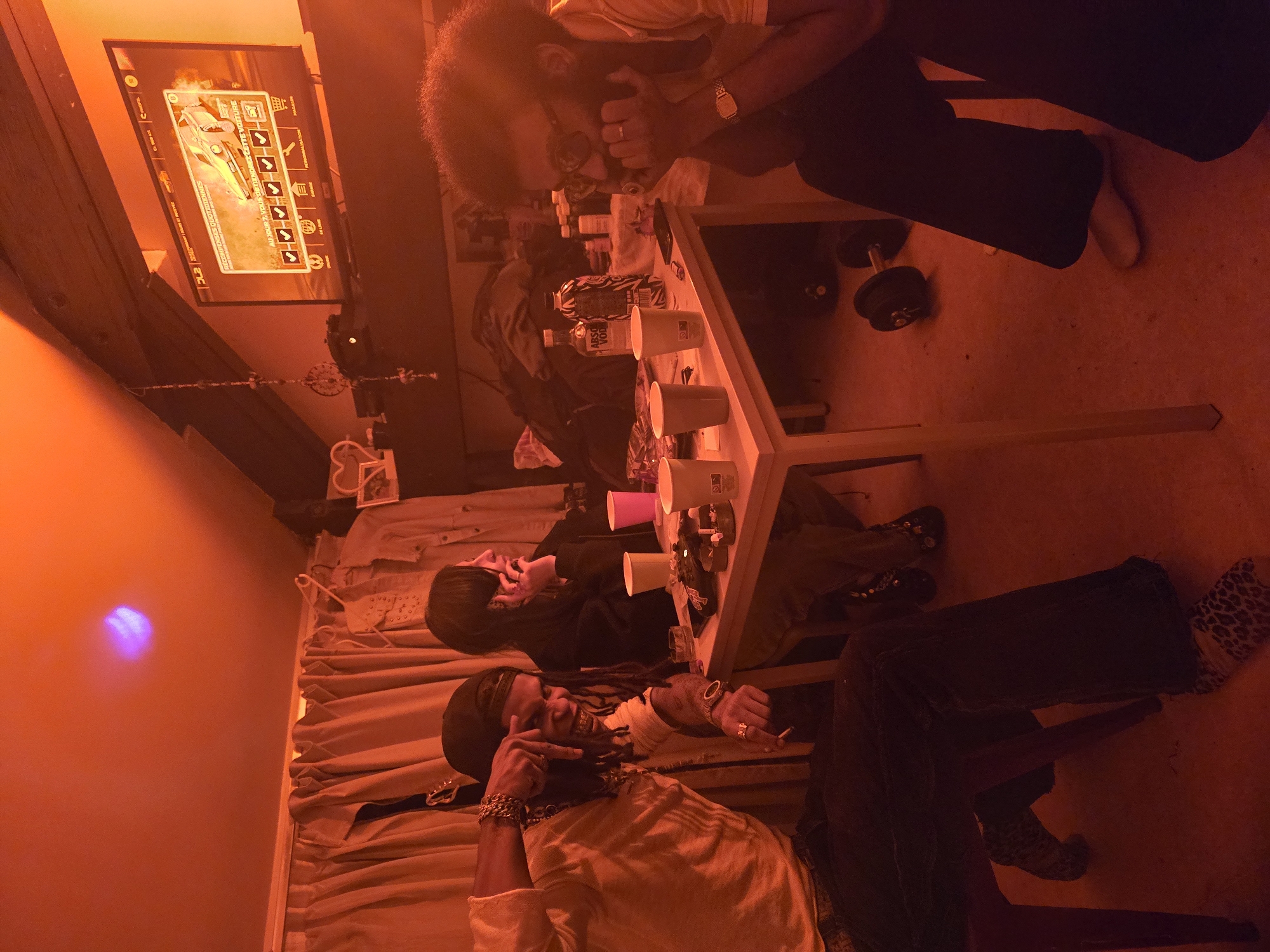 Three people are gathered around a table in a room with orange lighting. Two men are seated on either side of the table, while a third stands at the head. The table is covered in various items including cups, bottles, and other objects. A TV is mounted on the wall, displaying a menu or interface. The room appears to be a living space, with curtains and other furnishings visible in the background. There is no information to confirm the location as Paris, France.