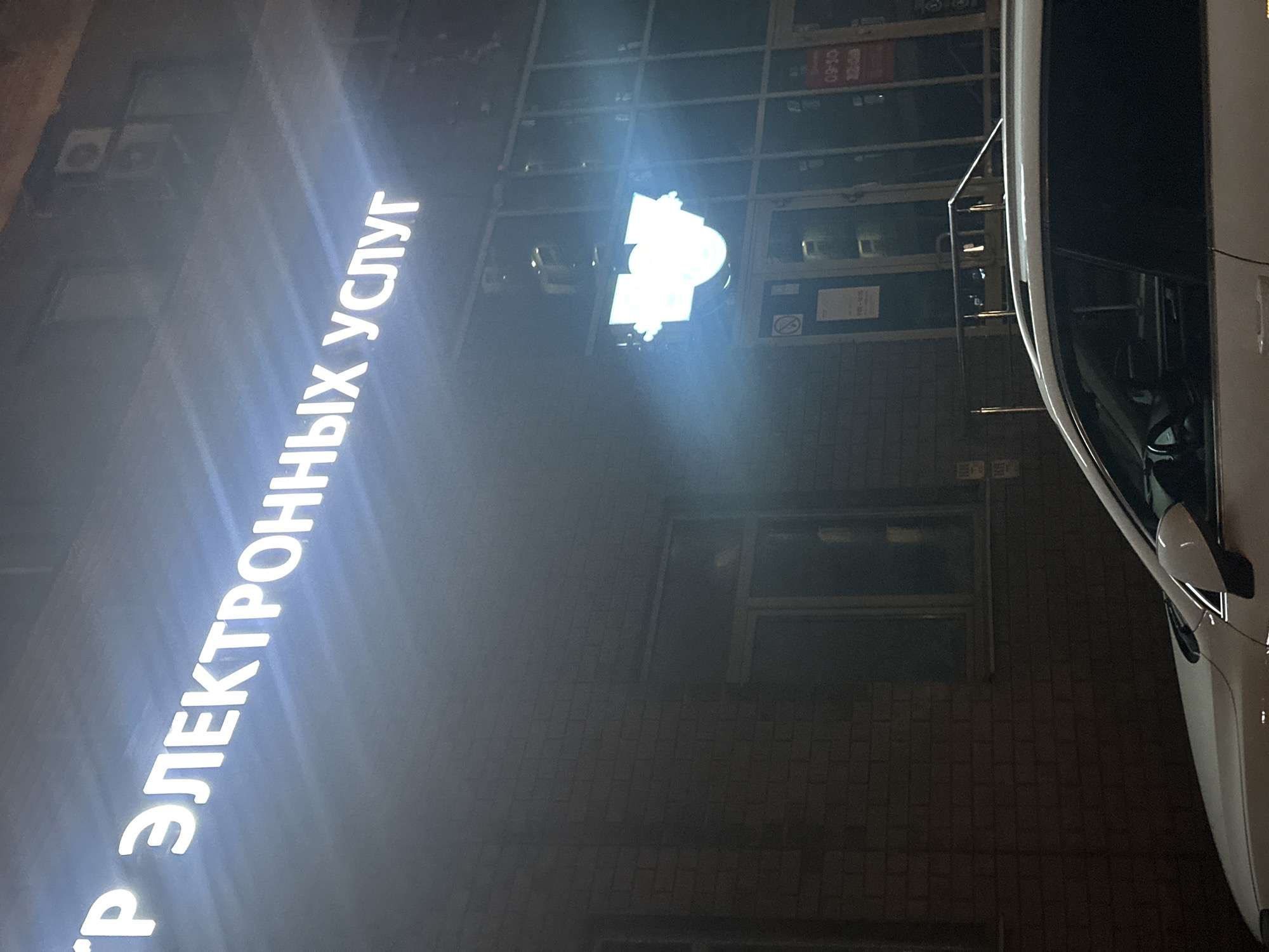 The image captures an exterior nighttime view of a building in Kazan, Russia, displaying the illuminated inscription "ЦЕНТР ЭЛЕКТРОННЫХ УСЛУГ" (Center of Electronic Services). The building's facade is constructed with a gray-bricked exterior with several windows, and a sign depicting an emblem above the entrance. Adjacent to the building, a white car is partially visible on the right side of the frame.