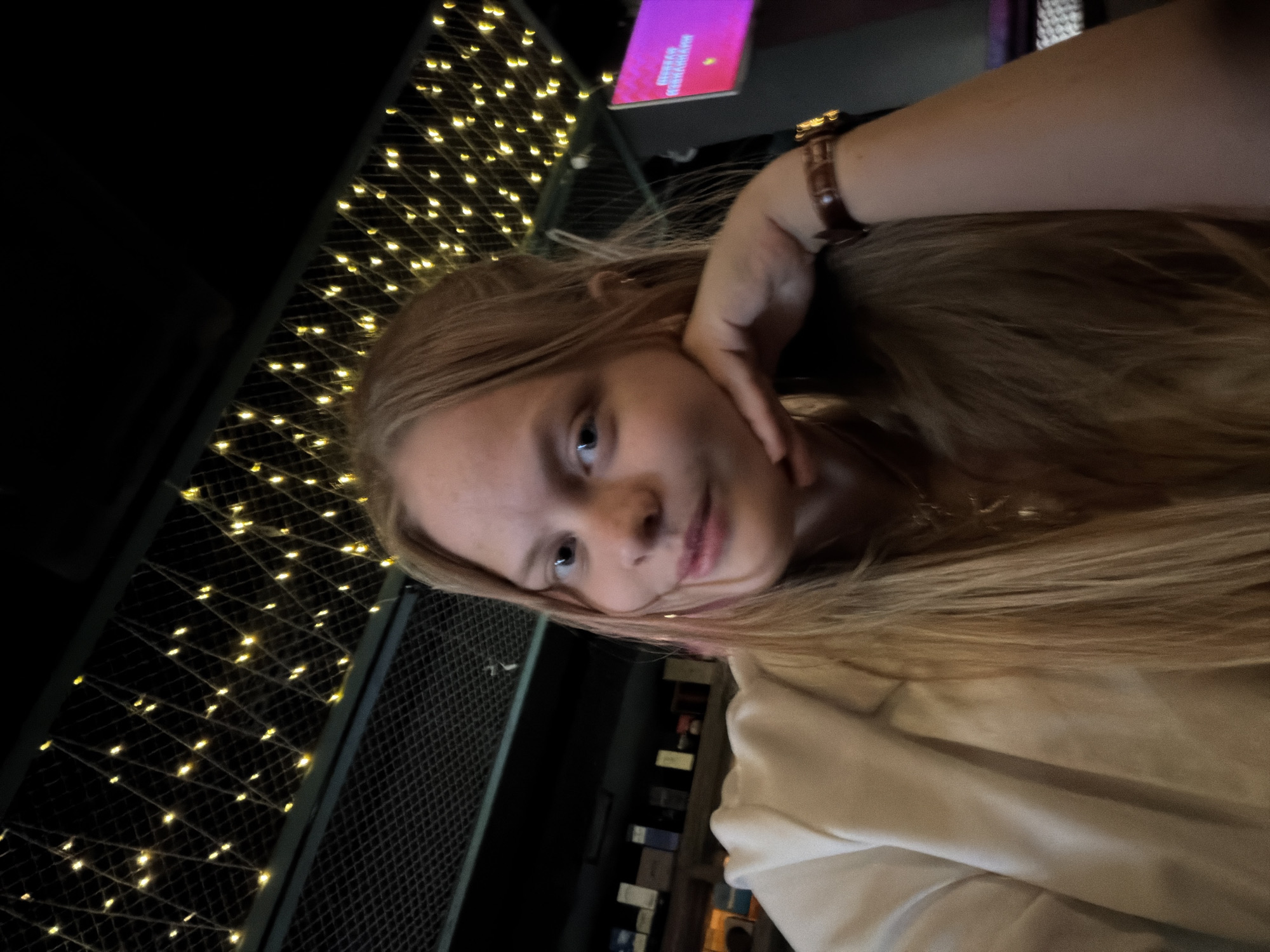 A young person with long blonde hair and blue eyes is posing for a selfie indoors. They are wearing a light-colored top and a wristwatch. The background features a dark interior with string lights hanging from a metal grid, and a blurred sign above. The location appears to be in Izhevsk, Russia.