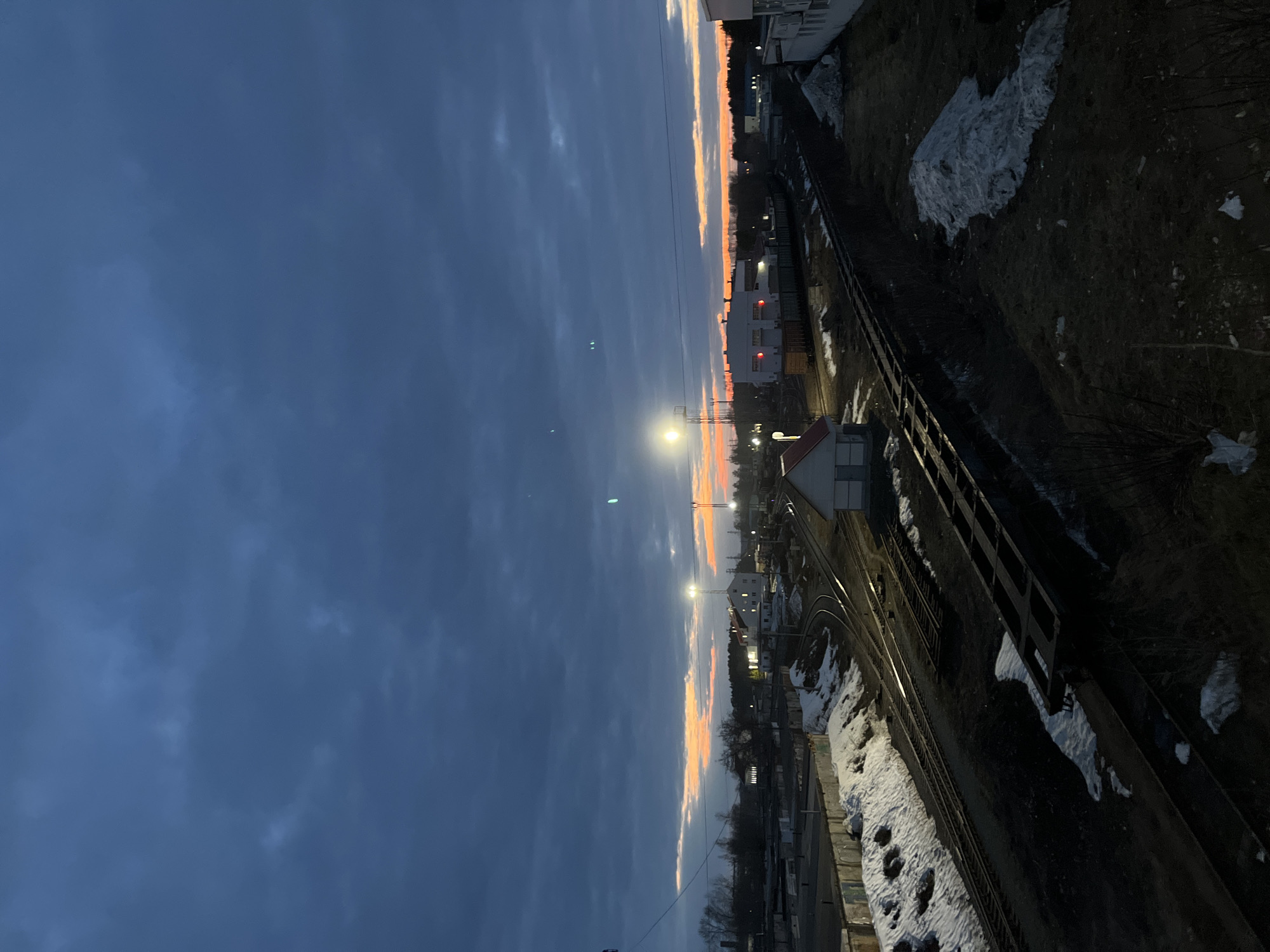 The image depicts a view of Chaikovsky, Russia at dusk. A railway track runs diagonally across the frame, with snow patches along its edge. Beyond the track, a street is visible with buildings and streetlights. The sky transitions from a deep blue to orange hues at the horizon, suggesting either sunrise or sunset. The scene appears deserted, with no visible people.