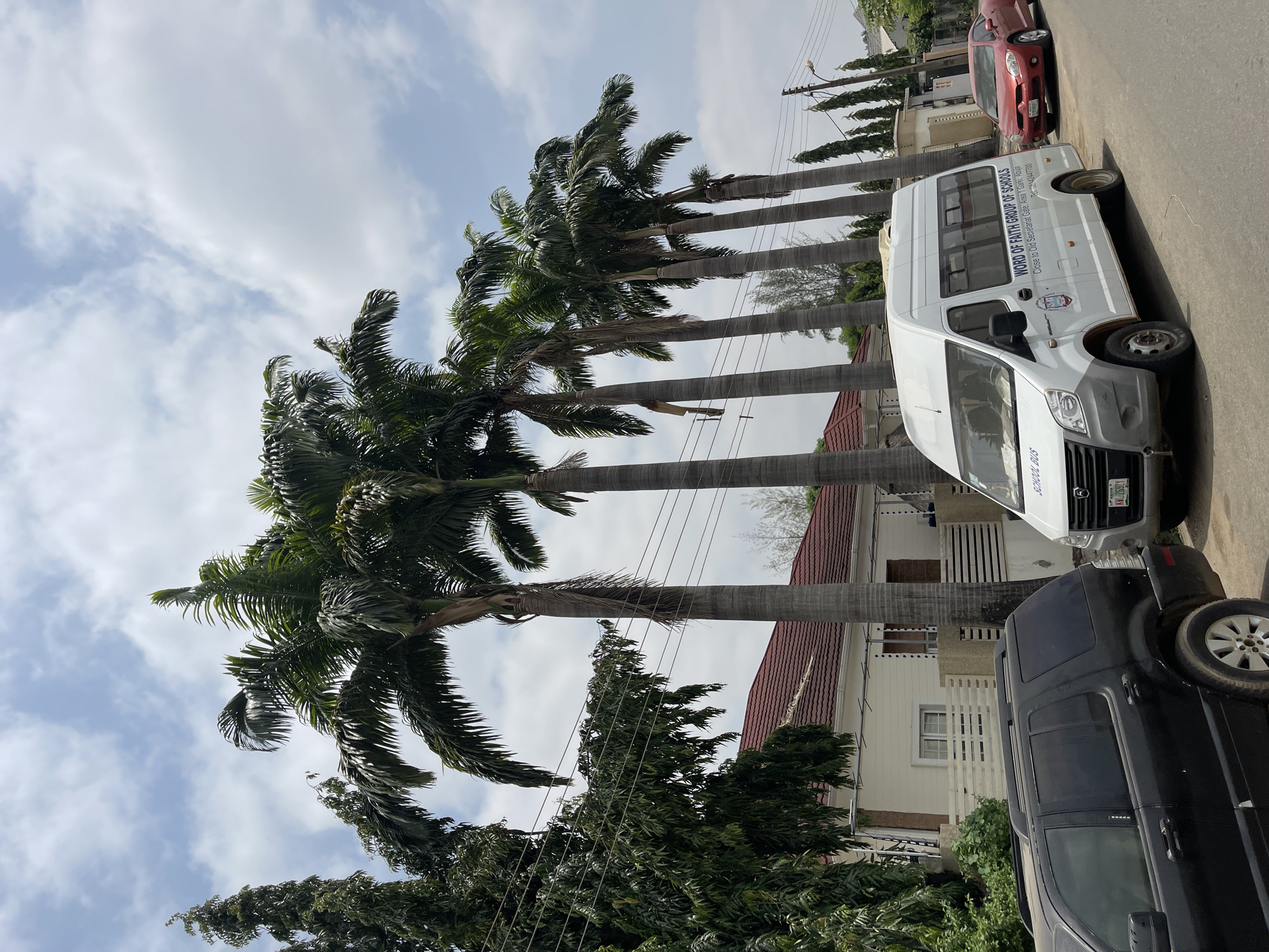 The image shows a street scene in Gwarinpa, Nigeria, featuring several tall palm trees lining the roadside. A white bus, identified as a "WORD OF FAITH GROUP OF SCHOOLS" school bus with license plate ML-269ZY, is parked along the street. A red car is parked further down the road, and a black SUV is parked closer to the foreground. The background includes residential buildings with red tile roofs. Power lines are visible running among the palm trees. The sky is partly cloudy.