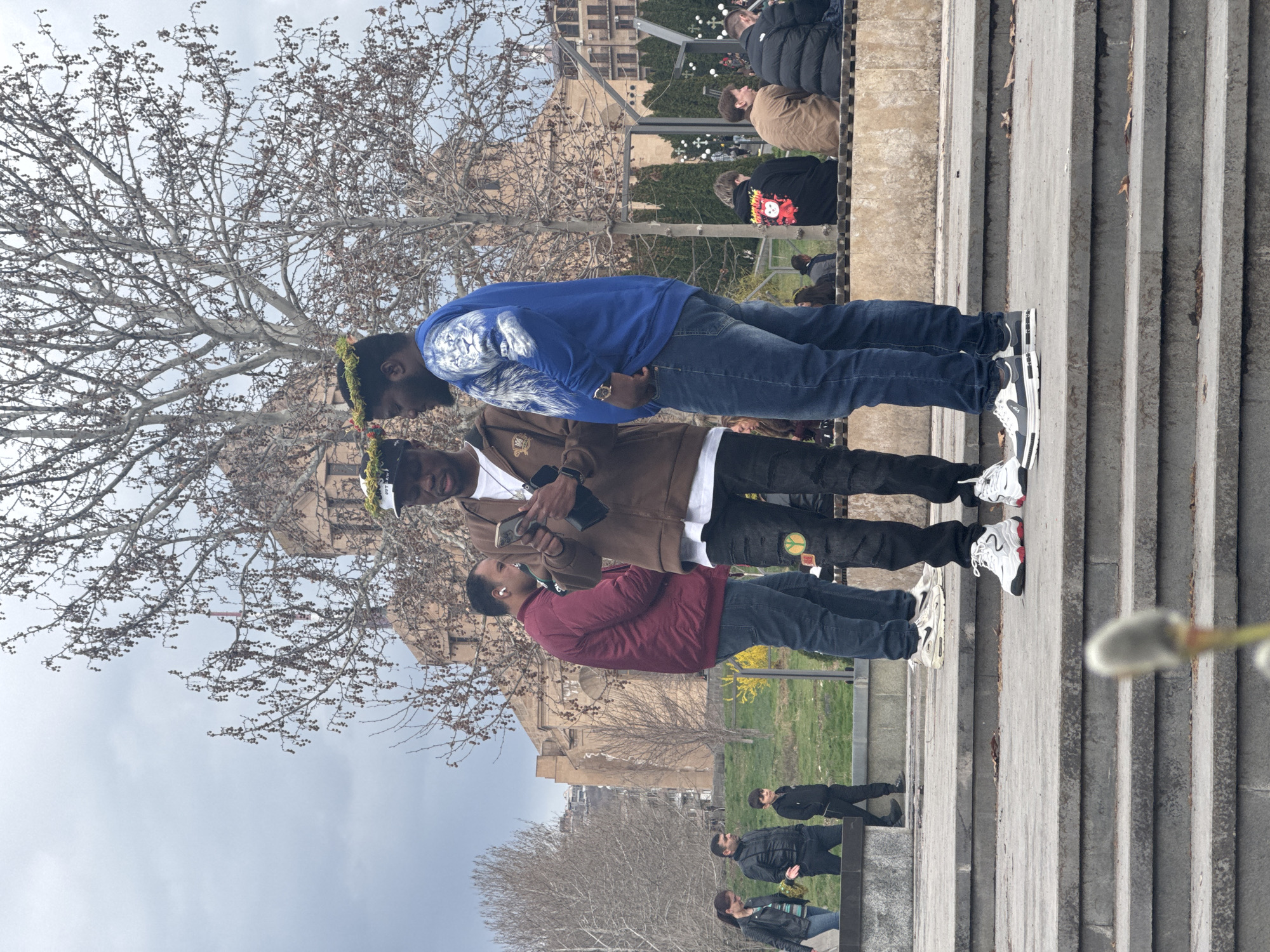 Three men stand in a vertical line on a concrete staircase. The first man is wearing a blue hooded sweatshirt and jeans. The second man is wearing a brown jacket over a white t-shirt and black jeans. The third man is wearing a maroon shirt and jeans. All three men are wearing sneakers. In the background, there is a building, trees, and other people. The photograph was taken in Yerevan, Armenia, based on the architectural style of the building in the background.