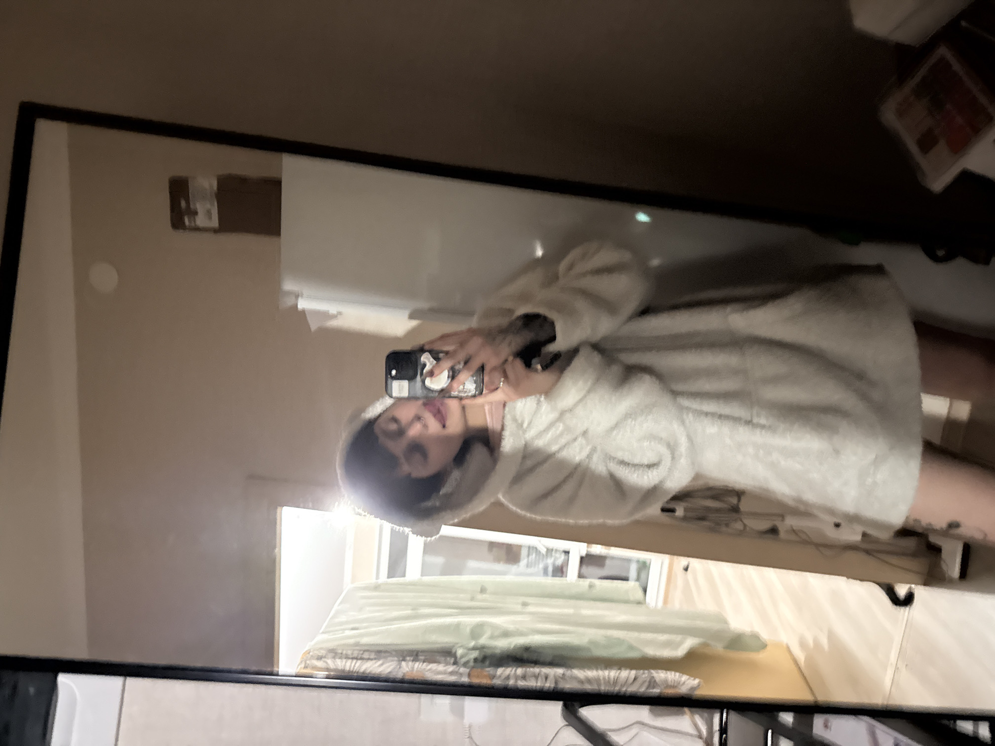 This image captures a young individual, likely female, with short dark hair and a septum piercing, taking a mirror selfie indoors in Saint Petersburg, Russia. The subject is wearing a light-colored, possibly cream or off-white, fuzzy hooded garment resembling a sweatshirt or tunic, featuring a large front pocket. Tattoos are visible on her left forearm/wrist and upper left thigh. She holds a dark smartphone with a light-colored accessory in her right hand, angled to capture her reflection, with her tongue slightly extended. The mirror, framed in black, reflects a room with a light-beige wall and a window or door with a white frame positioned in the background. To the left of the subject's reflection, several items of clothing, including a light green garment and a patterned one, hang on a visible rack. To the right of the subject, a white vertical object, possibly a refrigerator or cabinet, is reflected, topped by a brown box bearing a white label. The floor reflected in the mirror is light-colored. The wall immediately adjacent to the mirror on the right is a darker brown. A bright light source positioned behind or above the subject creates a halo effect around her head.