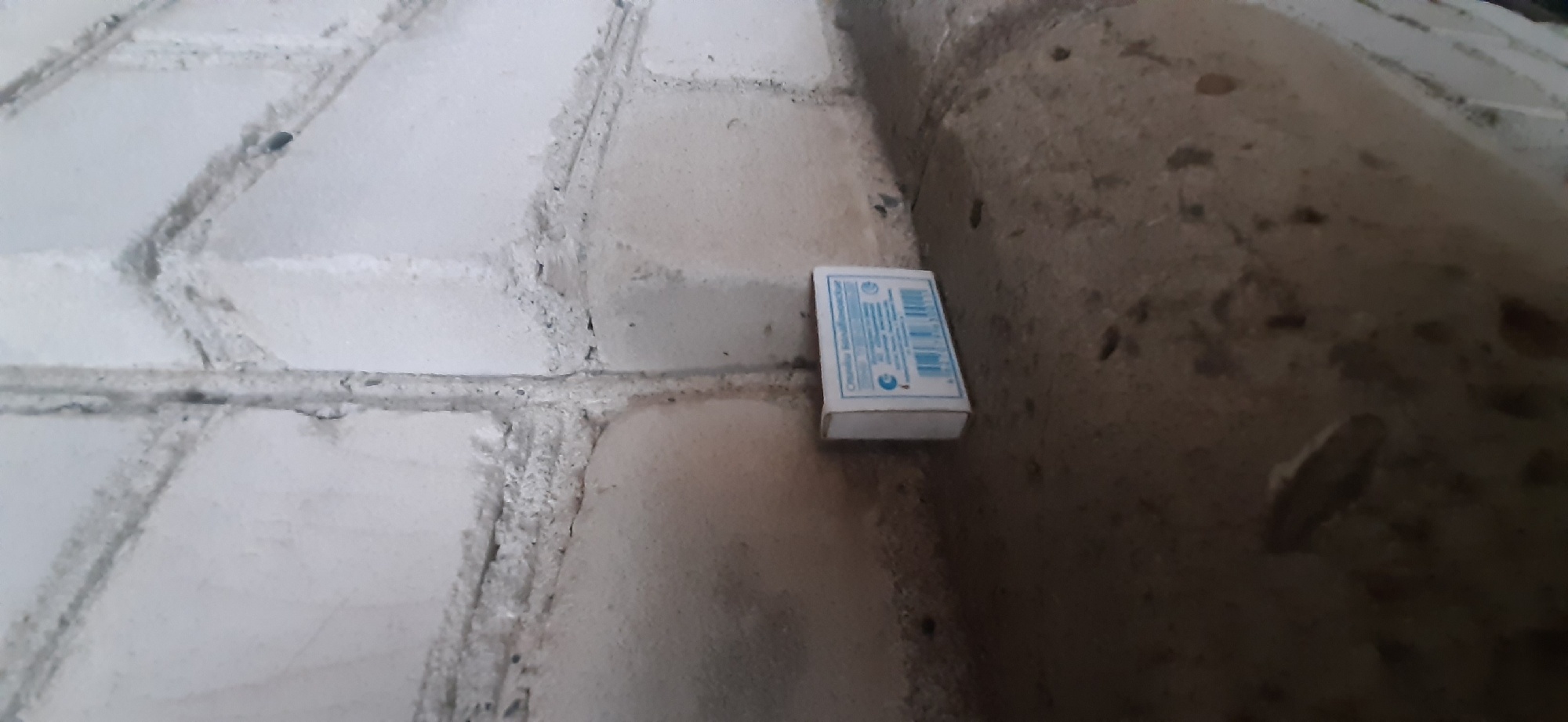 A white matchbox with blue lettering and a barcode is positioned on a concrete ledge. The background consists of a rough, white brick wall with gray grout lines. The lighting suggests an indoor or shaded outdoor environment.