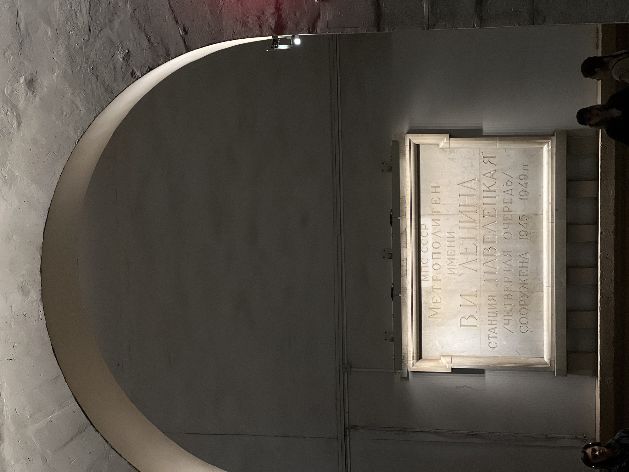 This image shows a memorial plaque located in the Moscow Metro system. The plaque is dedicated to the "Paveletskaya" station, which is named after V.I. Lenin. The plaque indicates that the station was part of the fourth construction phase (or line) and was built between 1945 and 1949. The text is written in Russian. The plaque is positioned on a wall and is framed by a border. The wall is visible through a large, arched opening, suggesting the plaque is mounted at the entrance to a part of the station. Several people are visible in the background, suggesting the area is accessible to the public.