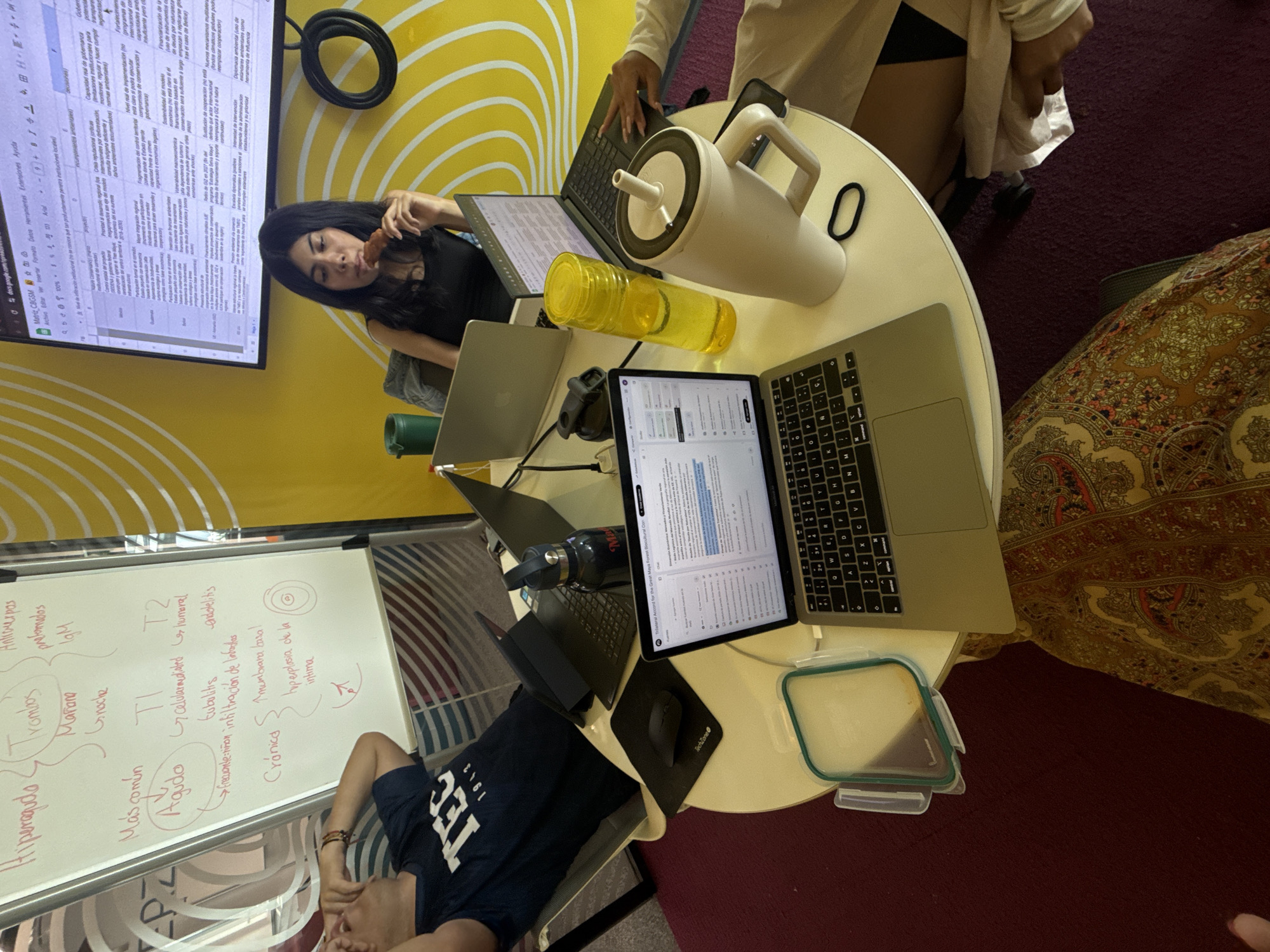 This media file captures an indoor study or work session with three individuals in what appears to be a collaborative workspace. A young woman with long dark hair, wearing a black top, is eating a snack while looking towards the camera. Another person, partially visible in a white top, is typing on a laptop at the table. A man, wearing a blue t-shirt with "TEC 1943" printed on it, is lying down on a chair or bench next to a whiteboard.
The circular table is filled with work essentials: multiple laptops, a yellow water bottle, a large white thermos, and a black water bottle. A computer mouse rests on a "TechZone" mousepad, and a food container is visible. One laptop screen displays a document titled "Trilateral Accord for the Great Maya Forest Biocultural Con..." (likely Conservation). A large monitor mounted on a bright yellow wall with white wavy patterns shows a Google Sheets spreadsheet titled "Matriz CBGSM." The spreadsheet contains extensive text in Spanish covering topics like project governance, funding, sustainability, and environmental diplomacy, with phrases such as "Capacidad real de gobernanza" and "Diplomacia ambiental."
Behind the man, a whiteboard displays handwritten notes in red marker, primarily in Spanish. These notes appear to be medical or biological terms, including "Agudo" (Acute), "Crónica" (Chronic), "T1," "T2," "IgM," "Celularmediated," "Humoral," "Tubulitis," "endotelitis," and "hyperplasia de la intima." The floor is covered in a dark red carpet, and a patterned fabric is visible on the right side of the frame. The overall scene suggests a focused yet relaxed academic or professional setting, potentially at a university, given the "TEC 1943" text on the t-shirt, possibly referring to Tecnológico de Monterrey.