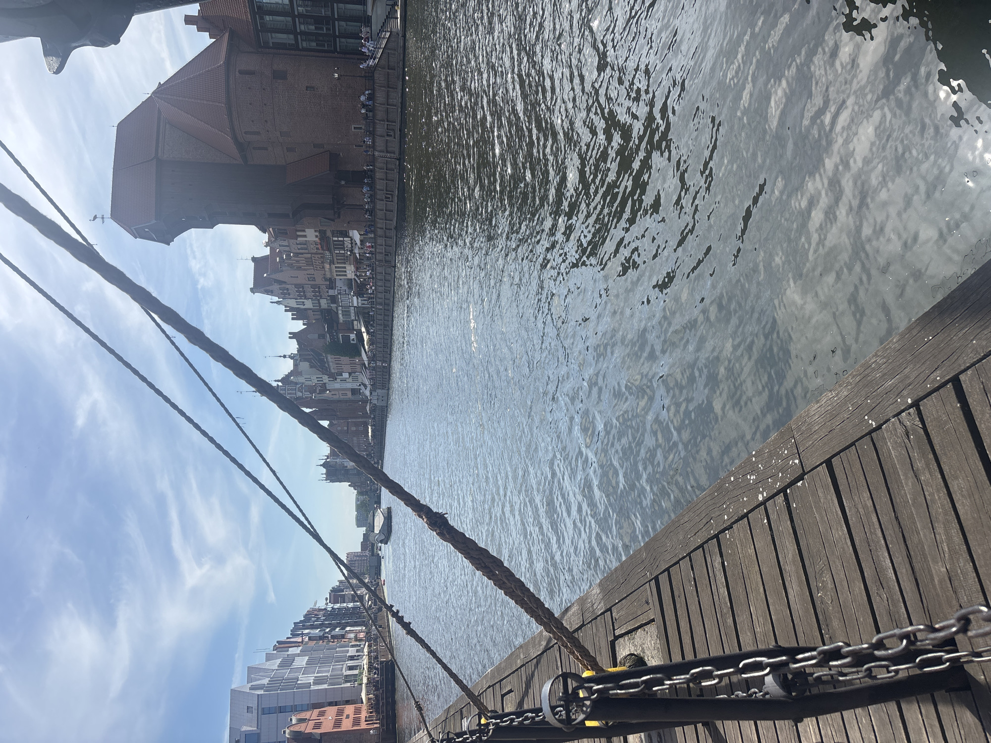 The image depicts a waterfront scene with buildings lining the river on the left side and a wooden pier on the right. The sky is blue with wispy white clouds, suggesting a clear and pleasant day, likely during daylight hours. The water is calm with ripples reflecting the sunlight.
Prominent structures include a large, historic brick building with a red-tiled roof, possibly a crane or warehouse, situated near the riverbank. Further along the waterfront, a line of multi-story buildings with varying architectural styles, some modern and some older, creates a cityscape. On the pier, thick ropes are secured, and metal chains are visible, hinting at maritime activities. A few people can be seen on the waterfront promenade, suggesting a public area. The perspective is from the pier, looking towards the river and the opposite bank. The overall atmosphere appears peaceful and scenic, indicative of a tourist or urban waterfront. There is no visible text in the image.