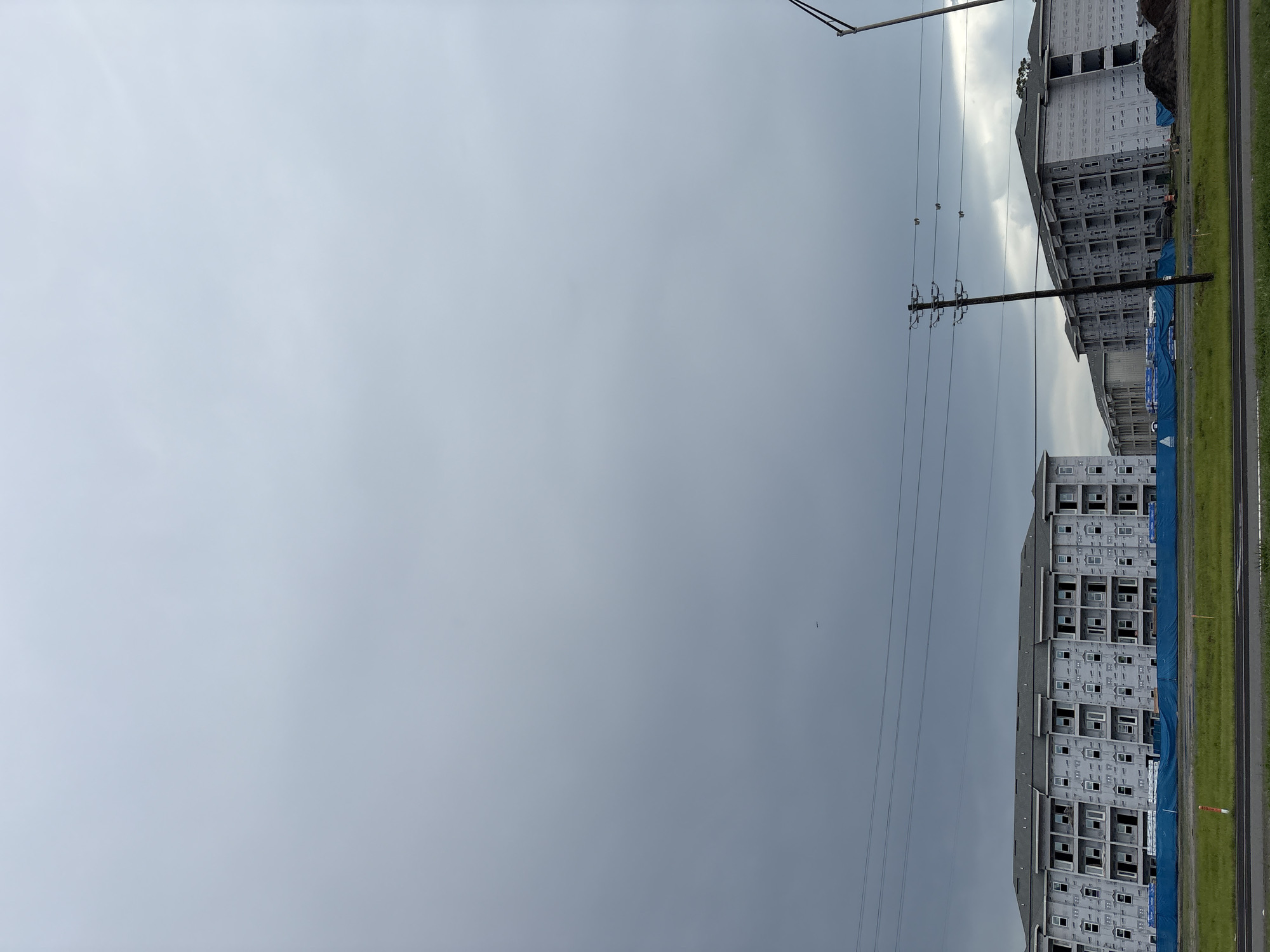 The image, taken in Bradenton, United States, depicts an overcast sky with dense clouds creating a somber atmosphere. In the foreground, a row of multi-story apartment buildings is under construction. These buildings are clad in a light-colored material, possibly house wrap, and many windows are visible. Blue tarps are spread across the ground in front of the buildings, likely covering materials or protecting the site.
Several utility poles with power lines traverse the scene, some appearing old and others more modern. To the right, a tall, dark pole with a light fixture is visible. The ground in front of the construction site is covered in green grass. In the distance, more buildings can be seen, also in various stages of construction, extending the scope of the development. A small dark object, possibly a bird or a drone, is visible in the sky. The overall impression is of a large-scale residential construction project in progress under a heavy, cloudy sky.