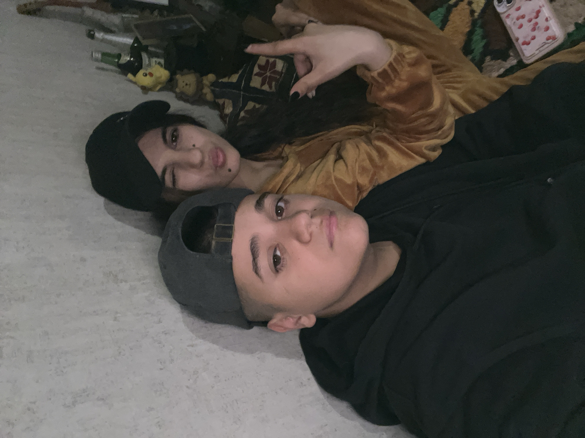 Two individuals are lying down, indoors. The person in the foreground is wearing a gray baseball cap and a black hooded sweatshirt. Behind them, the other person wears a black beanie, a mustard-colored shirt, and makes a hand gesture with black nail polish. Several bottles and small plush toys are in the background. A cherry-patterned phone case rests on a patterned pillow. The scene is captured from an overhead angle. No evidence suggests the location is Lozarevo, Bulgaria.