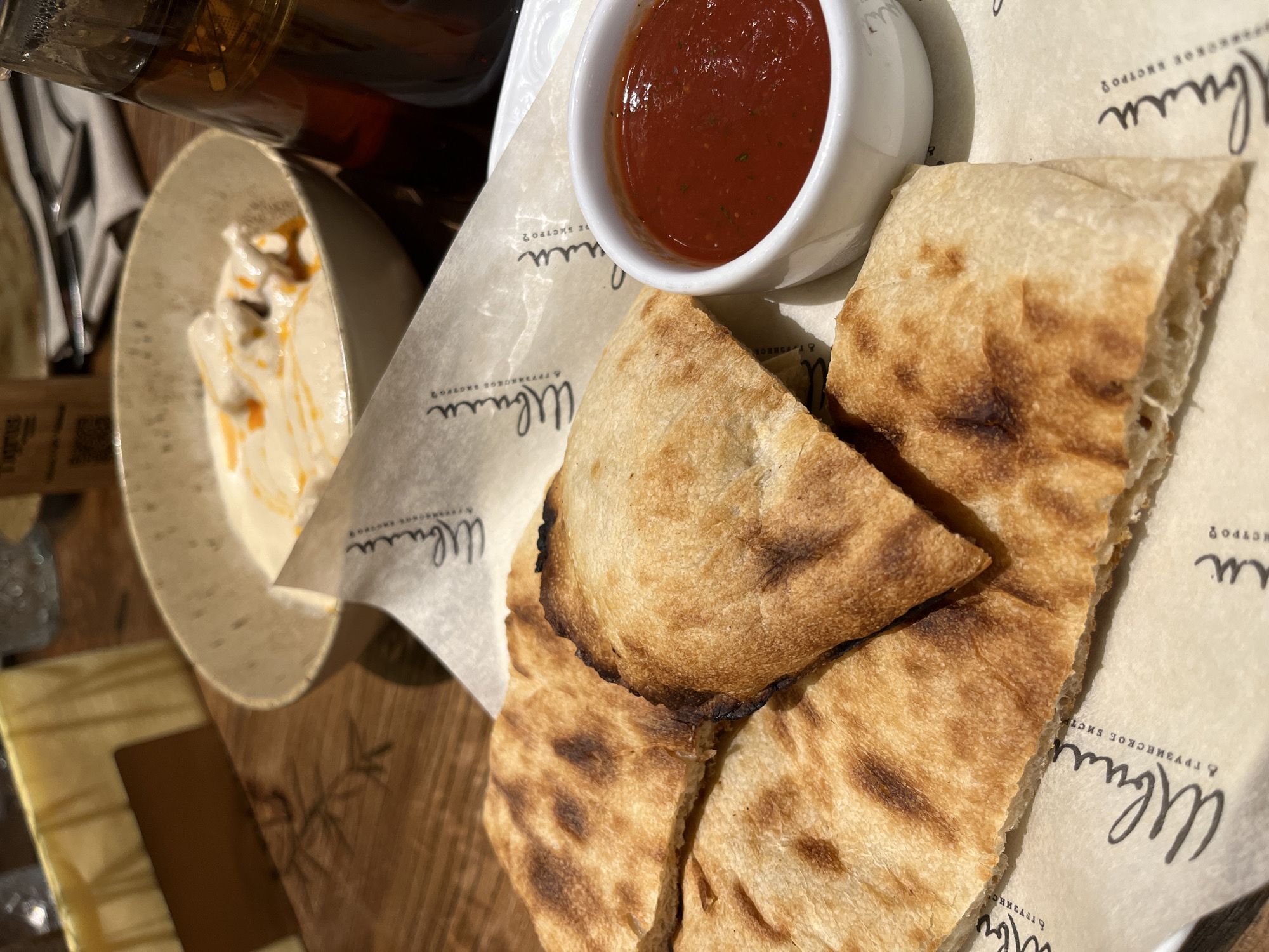 In the city of Khimki, Russia, a close-up, eye-level, indoor shot focuses on a dish of flatbread with a side of red sauce. The flatbread, light brown with darker toasted areas, is presented on a piece of paper with a script logo. The sauce is in a small white bowl. In the background, a larger bowl contains a creamy dish with what appears to be pieces of chicken and a drizzle of orange oil. A glass carafe containing a dark amber liquid is visible to the right, and a block of yellow cheese is on the left. The setting appears to be a restaurant, with wooden table surfaces visible.