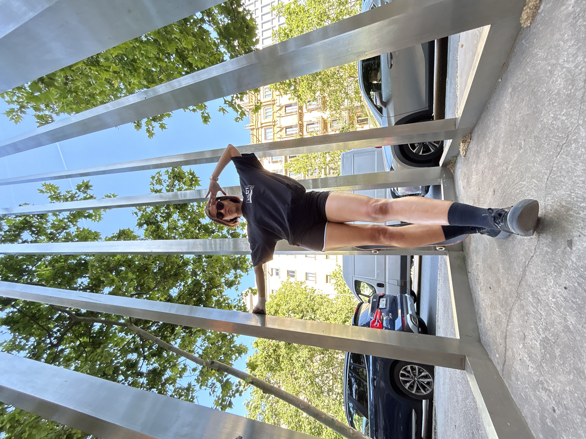 A full-body shot shows a woman posing in front of a modern metal structure on a sunny day in Barcelona, Spain. The woman is wearing a black t-shirt, black shorts, sunglasses, and black athletic shoes with socks. She is holding her arm above her head and leaning slightly on the metal structure. The structure consists of multiple vertical metal bars, creating a fence-like appearance. Behind the structure, there are trees with green leaves and a building with multiple windows. Cars are parked on the street behind the structure. The ground is concrete. The sky is blue and clear. The photograph is taken from a low angle, accentuating the height of the structure.