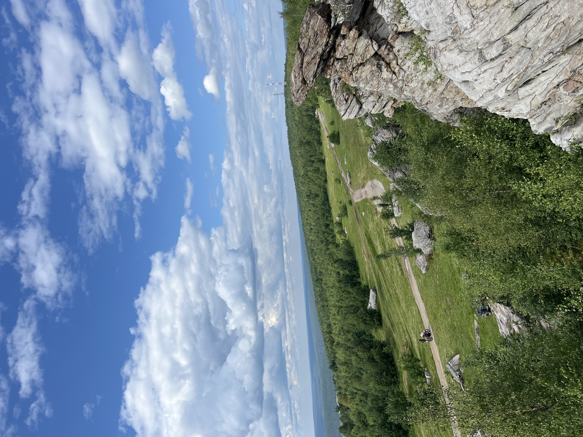 This image captures a scenic vista from a rocky overlook in Gubakha, Russia. In the foreground, large, weathered rock formations dominate the right side of the frame, their textured surfaces a mix of grays and browns. Lush green trees and bushes sprout from crevices and the surrounding terrain. A dirt path winds its way through a rolling, verdant hillside dotted with more boulders. In the midground, two individuals are visible on a dirt path, one on a bicycle and another on a motorcycle, suggesting recreational activity. Further in the distance, a dense forest stretches across the landscape, leading to a hazy horizon. Two tall transmission towers stand against the sky in the far distance. The sky above is a vibrant blue, punctuated by fluffy white cumulus clouds.