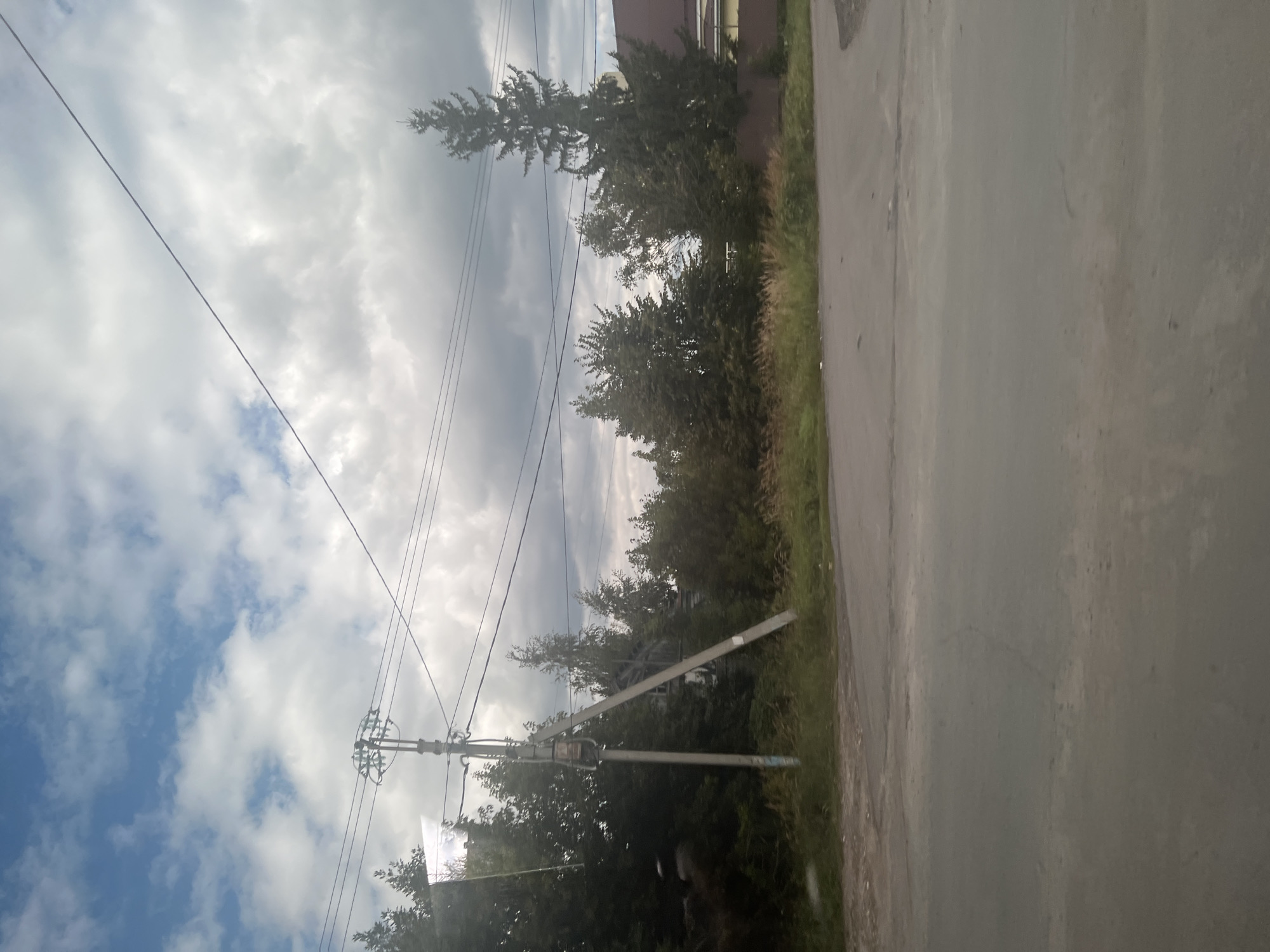 The image shows a street scene with a power line pole in the foreground. The pole is gray and has multiple wires extending from it. To the left of the pole, there are trees with dark green foliage. Behind the trees, a partially visible house with a brown roof and yellow siding can be seen. The sky is partly cloudy with blue sky peeking through. The ground in the foreground is an asphalt road. This scene appears to be in a residential area of Ulyanovsk, Russia, based on the architectural style of the house and the presence of overhead power lines.