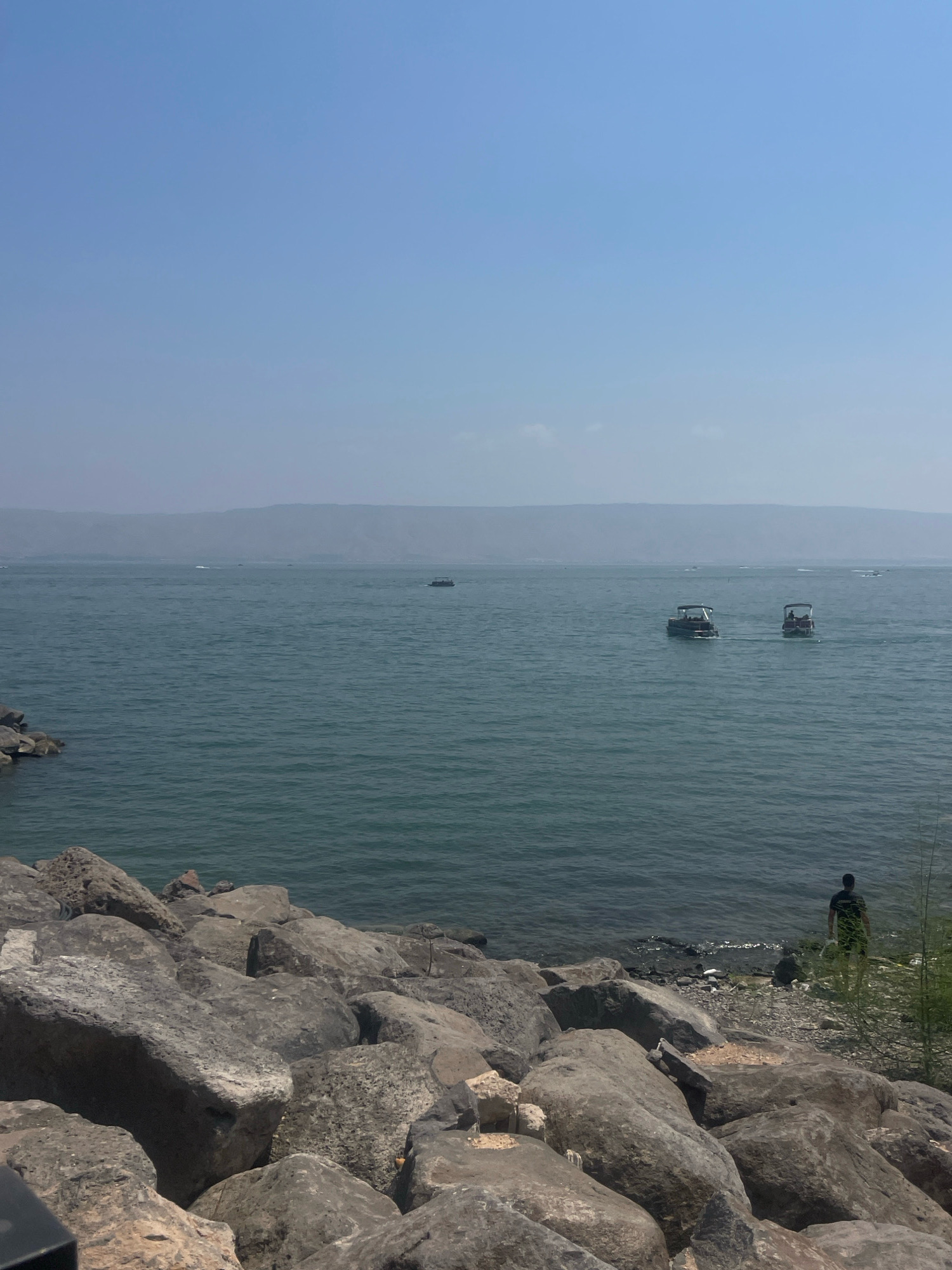 The image depicts a view of the Sea of Galilee in Tiberias, Israel. In the foreground, a rocky shoreline is visible, with large, grey stones forming the edge of the land. The sea is a deep blue-green, with gentle waves. Several boats are scattered across the water; two in the middle ground appear to be motorboats carrying passengers. In the distance, hazy hills form the horizon. On the right side of the frame, a lone individual stands on the shore, facing the water. The sky is a clear, pale blue.