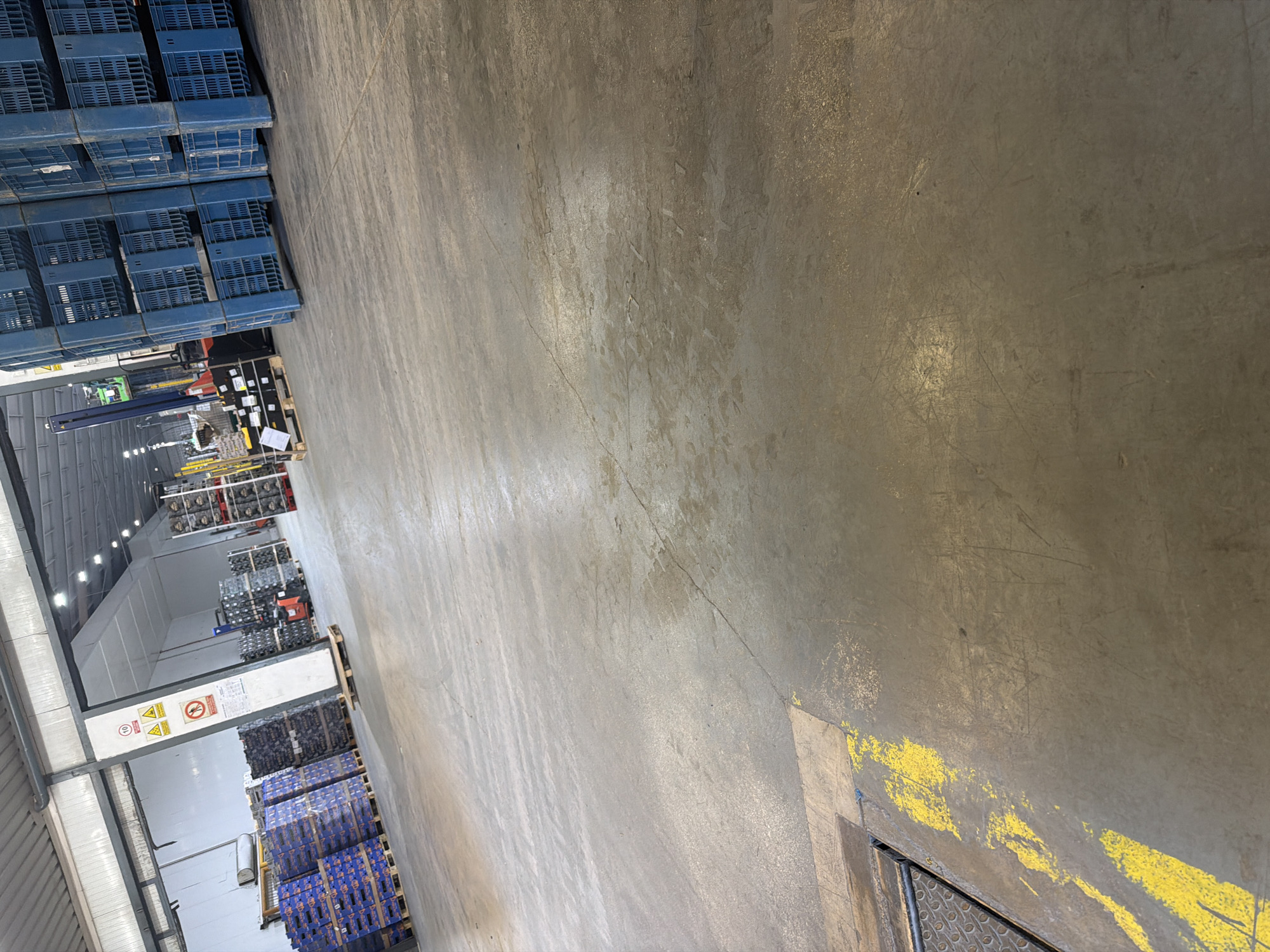 The image shows the interior of a warehouse, likely a distribution center or storage facility, located in Benifairó de la Valldigna, Spain. The floor is made of concrete and has yellow painted markings for safety or lane delineation. Large stacks of blue plastic crates are visible, some filled with products and others empty. In the background, there are loading docks and industrial equipment, including a forklift. The lighting appears to be artificial, typical of an indoor commercial space. The overall environment suggests an active operational facility for goods storage and movement.
