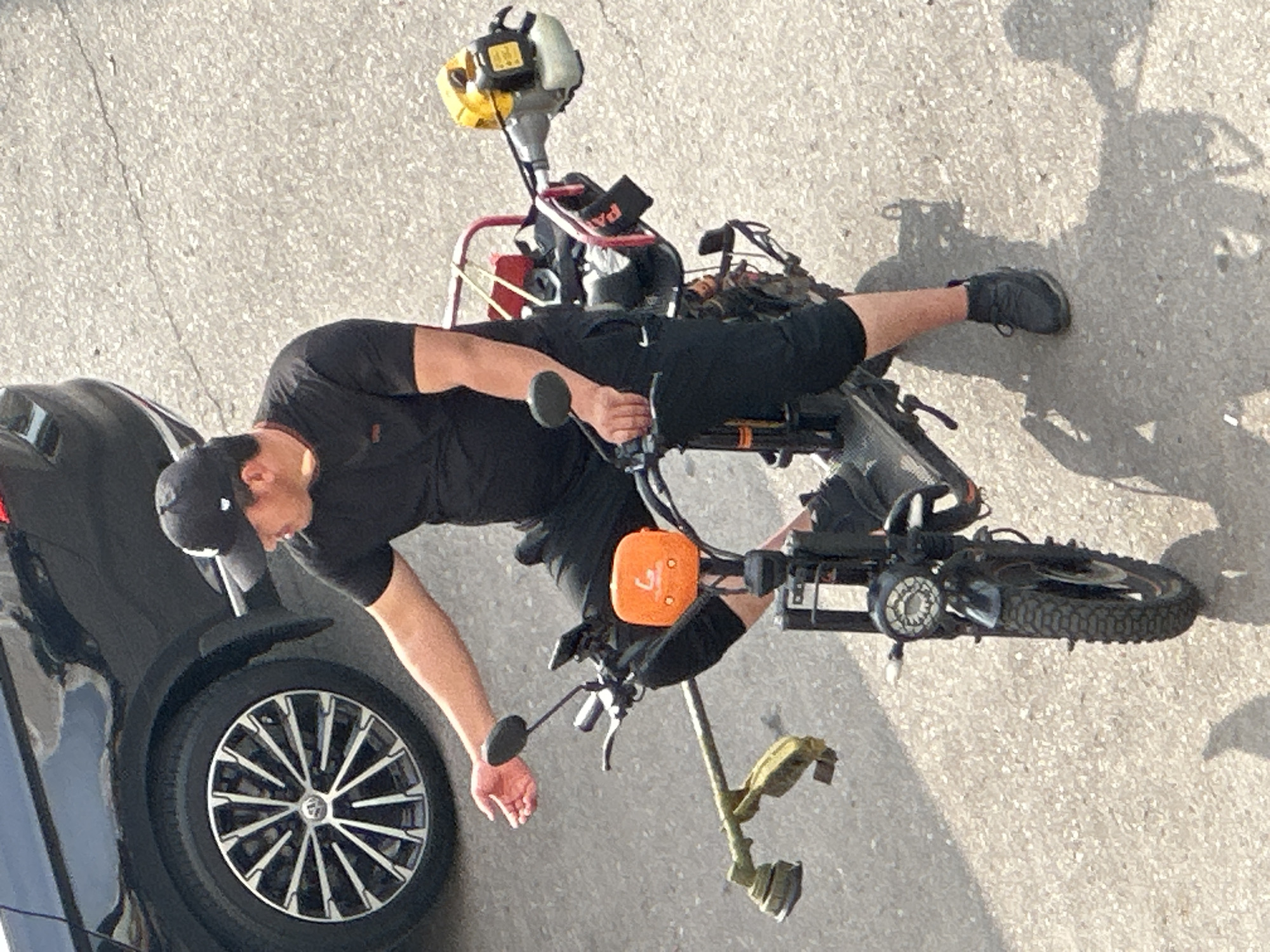 The image shows a man wearing a black t-shirt, black shorts, a black baseball cap, and black shoes, sitting on a modified electric scooter. The scooter has been outfitted with a red rack on the back, on which a yellow and black string trimmer engine is mounted. Another string trimmer head is attached to the front of the scooter. The man is holding his left hand out, palm up, as if gesturing. He appears to be in motion. The scooter is on a paved surface, likely a road or sidewalk, with a black car partially visible in the upper left corner. The lighting suggests it is daytime, with strong shadows cast on the ground. The scene is an outdoor urban environment. The provided context indicates the location is Podolsk, Russia.