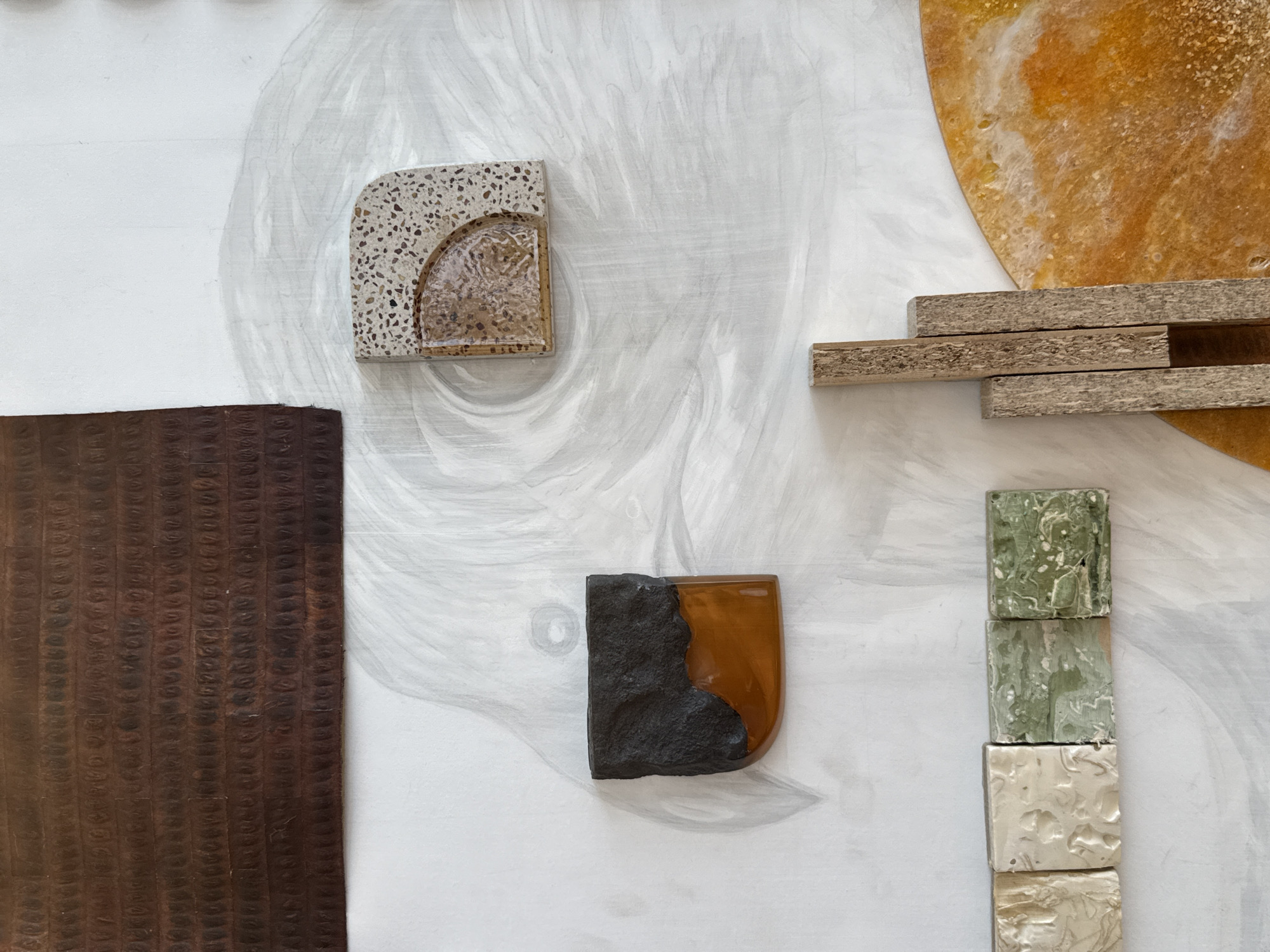The image shows a section of a wall with abstract decorative elements. The wall is white with swirling grey brushstrokes or textures. Attached to the wall are various materials and objects, including: a brown textured panel on the left; a square fragment with a speckled, terrazzo-like material forming a border around a resin-filled half-circle; a dark material paired with amber-colored resin; a stack of four small square tiles in shades of green and white; three wooden slats arranged horizontally; and a large yellow circle made of resin. The absence of any identifying architectural elements or specific landmarks means that the image cannot be directly tied to Dubai or any other specific location within the United Arab Emirates. The image is strictly a close-up of an interior design or art installation.