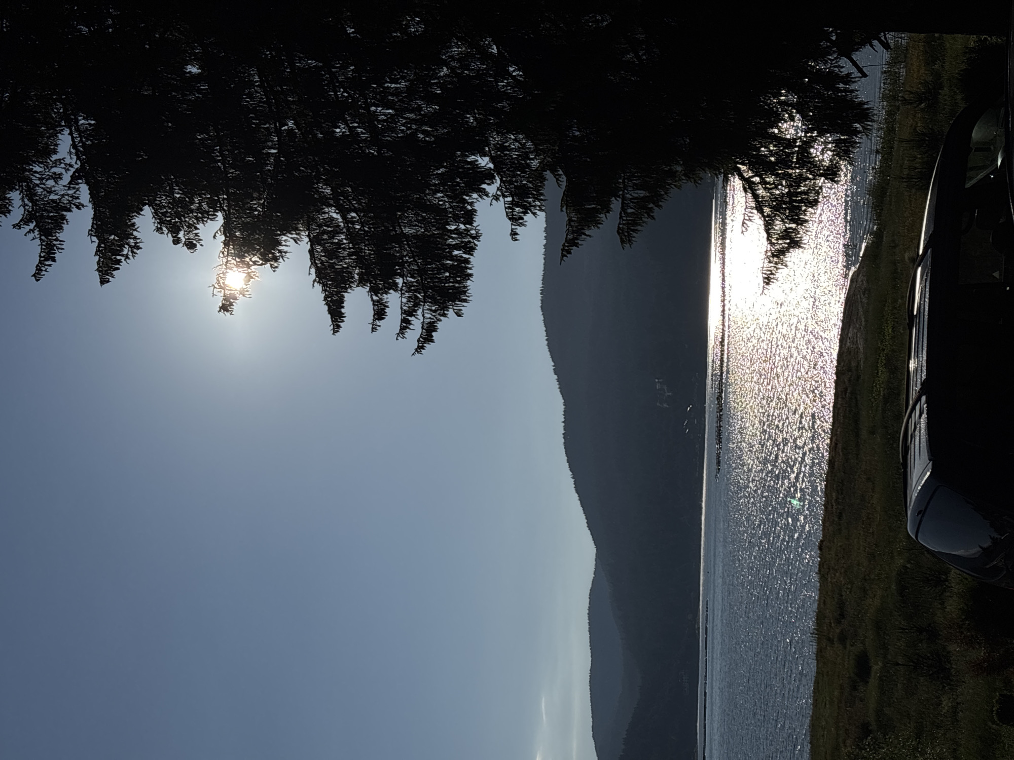 The image depicts a scenic outdoor environment with a body of water, rolling hills, and a large evergreen tree. The sun is shining brightly, creating a glare on the water. In the foreground, the roof and rear of a dark-colored SUV are visible, suggesting it is parked near the water. The scene is captured from a low angle, with the sun partially obscured by the tree's branches. The sky is clear and blue, and the overall impression is one of a peaceful, natural setting. The location is identified as Unknown, Unknown.