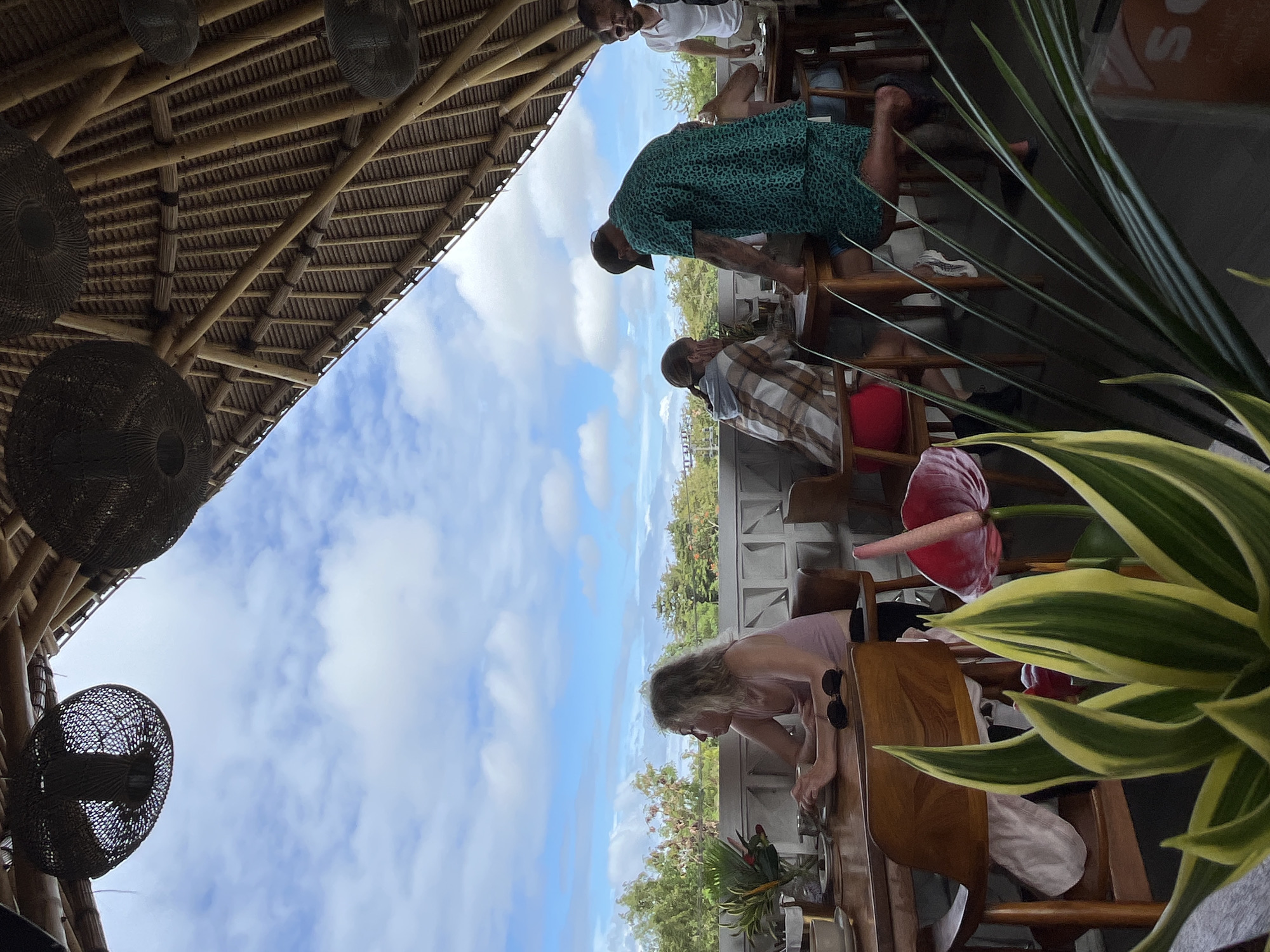 The image shows an outdoor dining area in Kuta Selatan, Indonesia. The scene is under a bamboo structure with hanging wicker lamps. Several people are leaning over a railing, looking out at the view. A woman in a pink top is at a table in the foreground. Potted plants with green and yellow leaves are also in the foreground. The background shows a blue sky with scattered clouds, trees, and buildings.