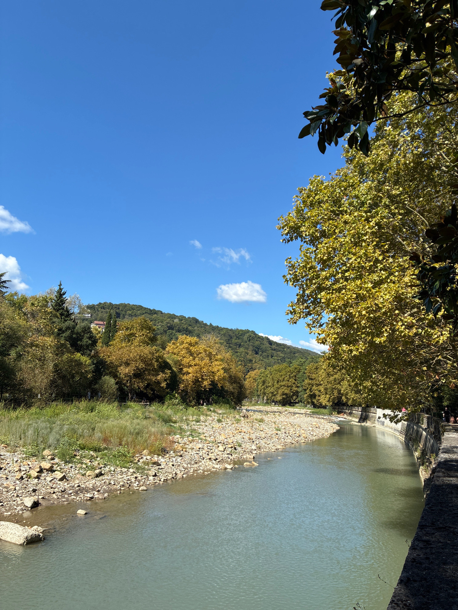The image depicts a scenic view of the Matsesta River in Sochi, Russia, on a clear day. The river flows gently, with its banks characterized by a mix of smooth stones and sparse vegetation. On the left side, the riverbank is rocky and appears somewhat dry, with patches of green grass. On the right side, a paved embankment with a stone wall runs along the river. Lush green trees with some autumn-colored foliage line the embankment, casting shadows on the path. In the background, rolling hills covered with dense green forest rise up to meet a bright blue sky dotted with a few white clouds. A building with a red roof is visible nestled among the trees on the hillside. A few people are seen walking along the embankment on the right side of the image, suggesting this is a public area. The overall atmosphere is tranquil and natural.
