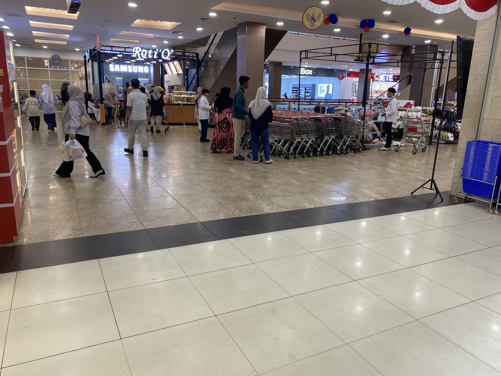The image depicts the interior of a shopping mall in Banda Aceh, Indonesia. The scene is dominated by a wide, tiled walkway with a darker tiled section marking a transition. Several individuals, including men, women, and children, are present. Some are walking, while others appear to be standing and conversing. In the background, various storefronts are visible, including one with a prominent "Roti 'O'" sign and another displaying "SAMSUNG." A long row of shopping carts is also a significant feature, suggesting a retail environment where customers use these for their purchases. The lighting appears to be artificial, typical of an indoor shopping space. Decorations, possibly for a festive occasion, are suspended from the ceiling. The overall atmosphere suggests a bustling public area within a commercial establishment.
