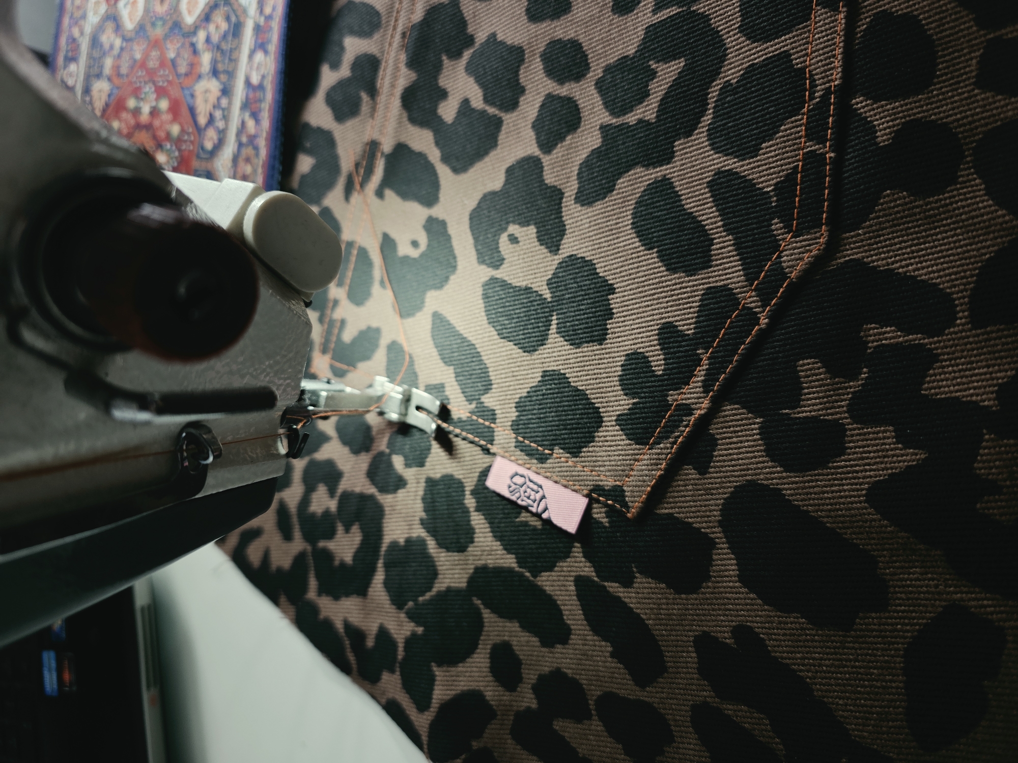 A close-up shot focuses on a sewing machine stitching leopard-print fabric. A small pink label with dark text is near the stitches. The sewing machine is silver and grey, with a needle and thread visible. The fabric has a light brown background with black leopard spots and orange stitching. In the background, a blurred image of what appears to be a rug with intricate patterns is visible. This image was not taken in St. Petersburg.
