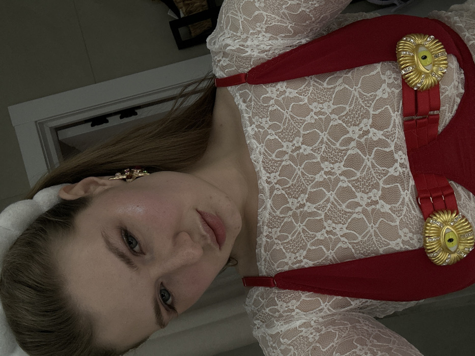 A woman in a white lace top and red suspender-like garment with gold eye-shaped decorations. She is fair-skinned with blonde hair, and wearing an earring on her right ear. The background features a white door and a dark-colored object.