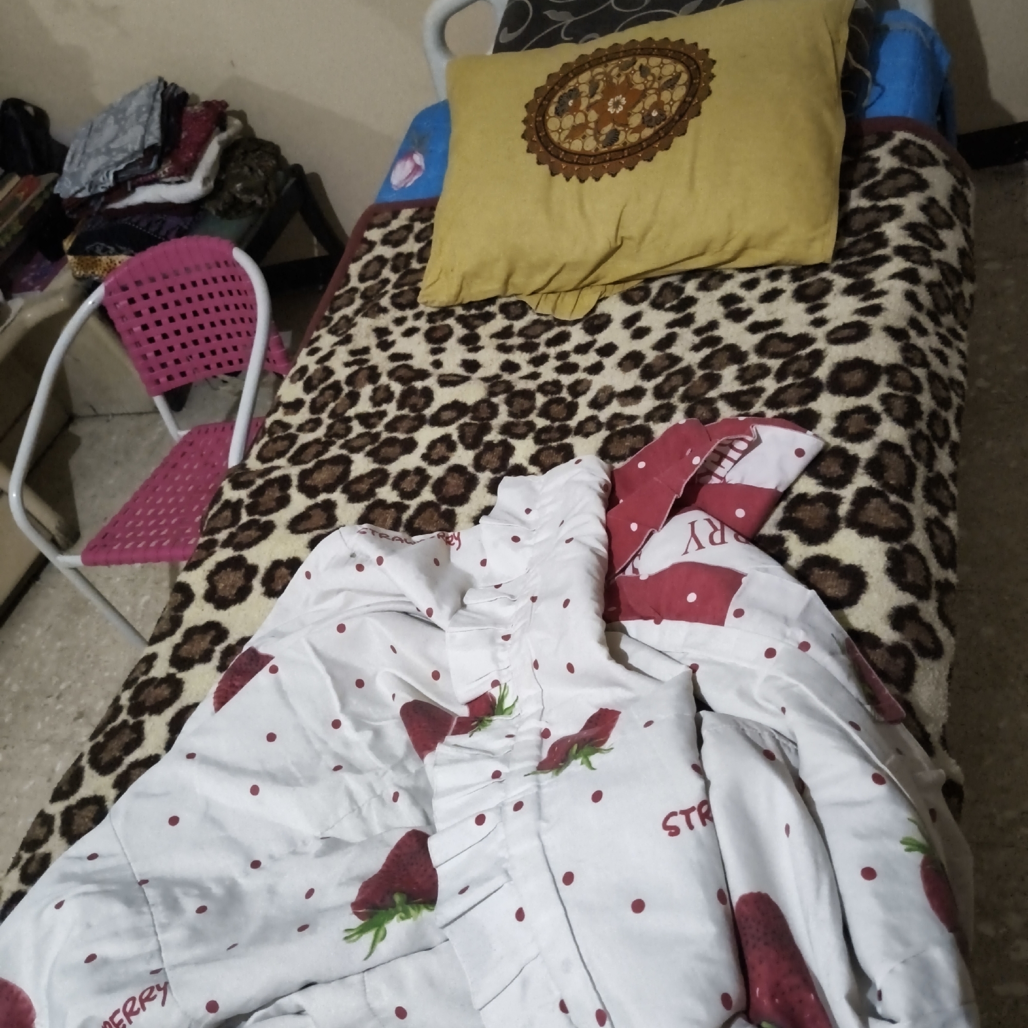 This is a photograph taken indoors, likely a bedroom, with a cluttered and lived-in appearance. The primary focus is a made-up bed with a leopard-print blanket covering the mattress. A yellow throw pillow with an embroidered brown and orange circular pattern rests on the bed. In the foreground, a white comforter with a strawberry pattern is folded and partially covers the bed. To the left of the bed, a pink plastic chair with a white metal frame is positioned. A pile of folded clothes sits on a dark-colored rack behind the chair. The lighting is dim, casting soft shadows throughout the room. The overall impression is one of a casual and somewhat disorganized personal space.