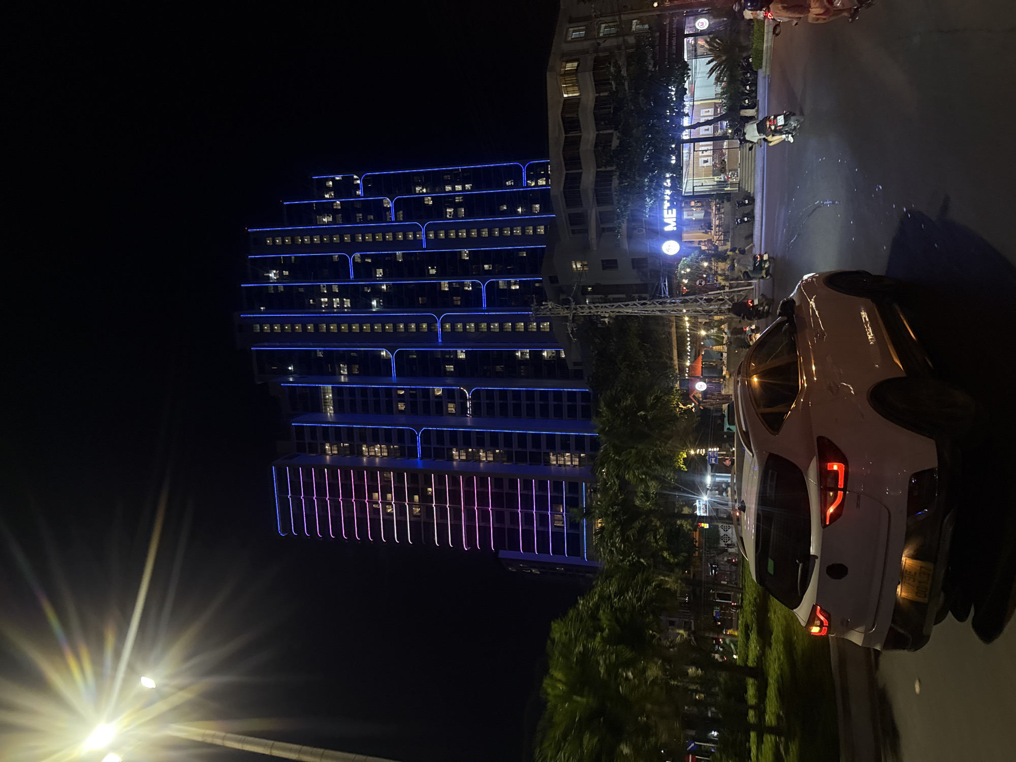 The image depicts a nighttime street scene in Bac Nha Trang Ward, Vietnam. A white SUV is in the foreground, driving away from the camera on a paved road. Behind the SUV, a tall, modern building is illuminated with blue and pink vertical lights. Palm trees and other greenery are visible on the left side of the road. Several motorcycles are also present on the road, with some riders visible. A building with a sign that reads "ME" is illuminated with bright lights in the background. The overall scene suggests a vibrant urban environment at night, with traffic and illuminated architecture.