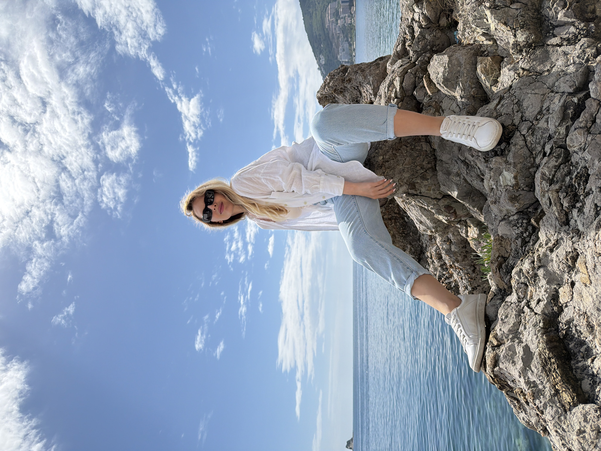 A woman with blonde hair wearing sunglasses, a white shirt, and light blue jeans, is perched on a large rocky outcrop overlooking the Adriatic Sea in Budva, Montenegro. She is wearing white sneakers and her limbs are positioned so that she is both sitting and standing on the rocks. The sky is blue with scattered white clouds. The rock formation is large and textured, with the sea stretching into the horizon behind it. Some buildings are faintly visible on a distant hillside.
