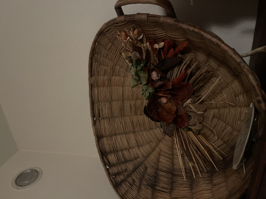 The image displays a large, oval-shaped woven basket, possibly used for decorative purposes, hanging against a light-colored wall in Dosso (Ciappanico), Italy. The basket is crafted from natural, light brown fibers with a distinct ribbed weaving pattern and features a sturdy handle at its top.

Inside the basket, an arrangement of dried botanical elements is prominently featured. This includes several large, deep reddish-brown dried flowers with visible stamens, intermingled with lighter-colored dried grasses, spiky seed heads, and smaller clusters of dried green and orange foliage. The overall impression of the arrangement is rustic, natural, and autumnal.

To the right side of the basket, a small, metallic or gray object, possibly a clip or a part of the basket's hanging mechanism, is partially visible. In the bottom left corner of the frame, a circular recessed light fixture is embedded in the ceiling or upper wall. The lighting is soft and ambient, indicating an indoor setting. There are no people visible, and no specific activity or event is taking place, making it a still life composition. No visible text is present in the image.