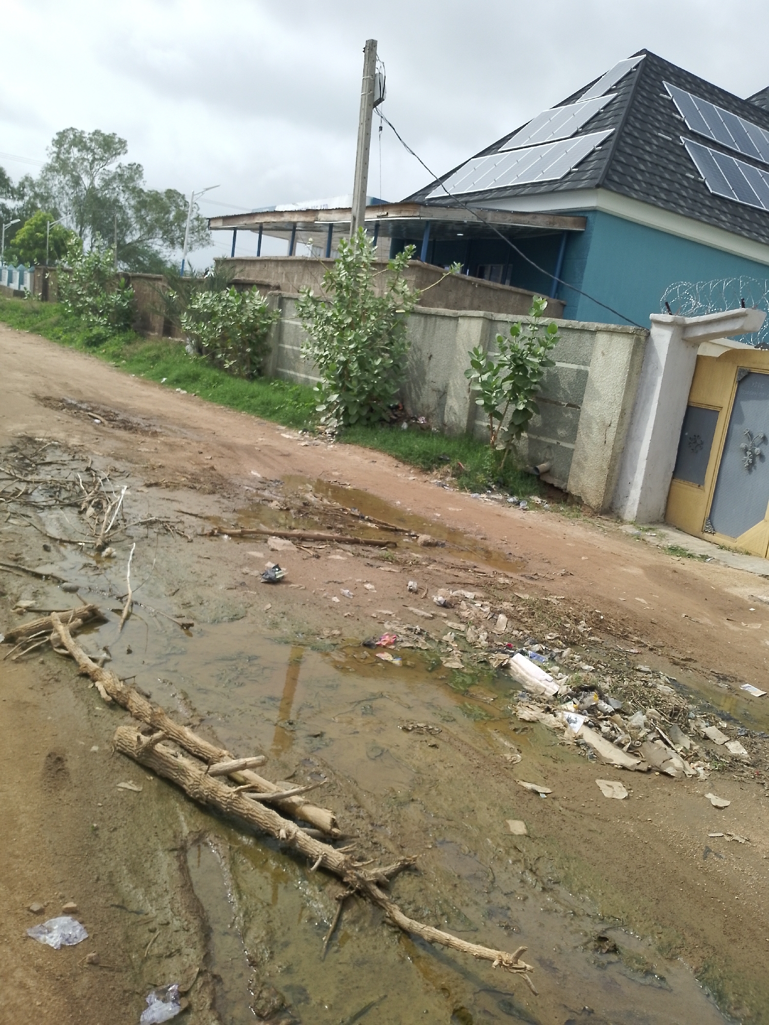 The image depicts a muddy dirt road with debris scattered across it. A pile of branches lies in the foreground, partially submerged in a puddle. In the background, a blue building with solar panels on its roof is visible, along with a concrete wall and a yellow gate. The sky is overcast.