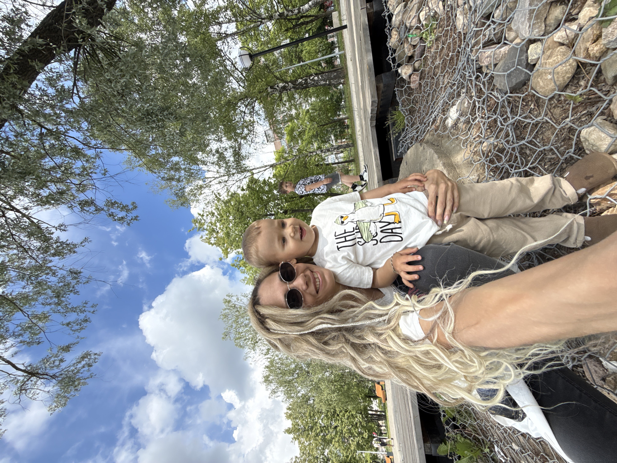 A rotated selfie-style image captures a cheerful woman holding a young child outdoors, likely in a park in Rusanovo, Russia. The woman, positioned closer to the camera, has long, wavy blonde hair and wears dark round sunglasses, smiling broadly. Her right arm, partially visible, suggests she is holding the camera. A young boy, likely a toddler, is held closely by the woman. He looks up with a wide smile, wearing a white t-shirt featuring a cartoon duck graphic and partially visible text "THE...DAY," along with light brown pants.
The background reveals a sunny day with a clear blue sky dotted with scattered white clouds. Numerous green trees with lush foliage, some appearing to be birch, are spread throughout the scene. A gabion-style wall, made of wire mesh filled with rocks, is prominently featured on the right side, alongside a wooden or concrete ledge. In the distance, another person is visible walking. The bright light suggests it is midday or early afternoon. The overall emotion conveyed is happiness and affection between the woman and child. The image is oriented 90 degrees clockwise from a standard portrait view.