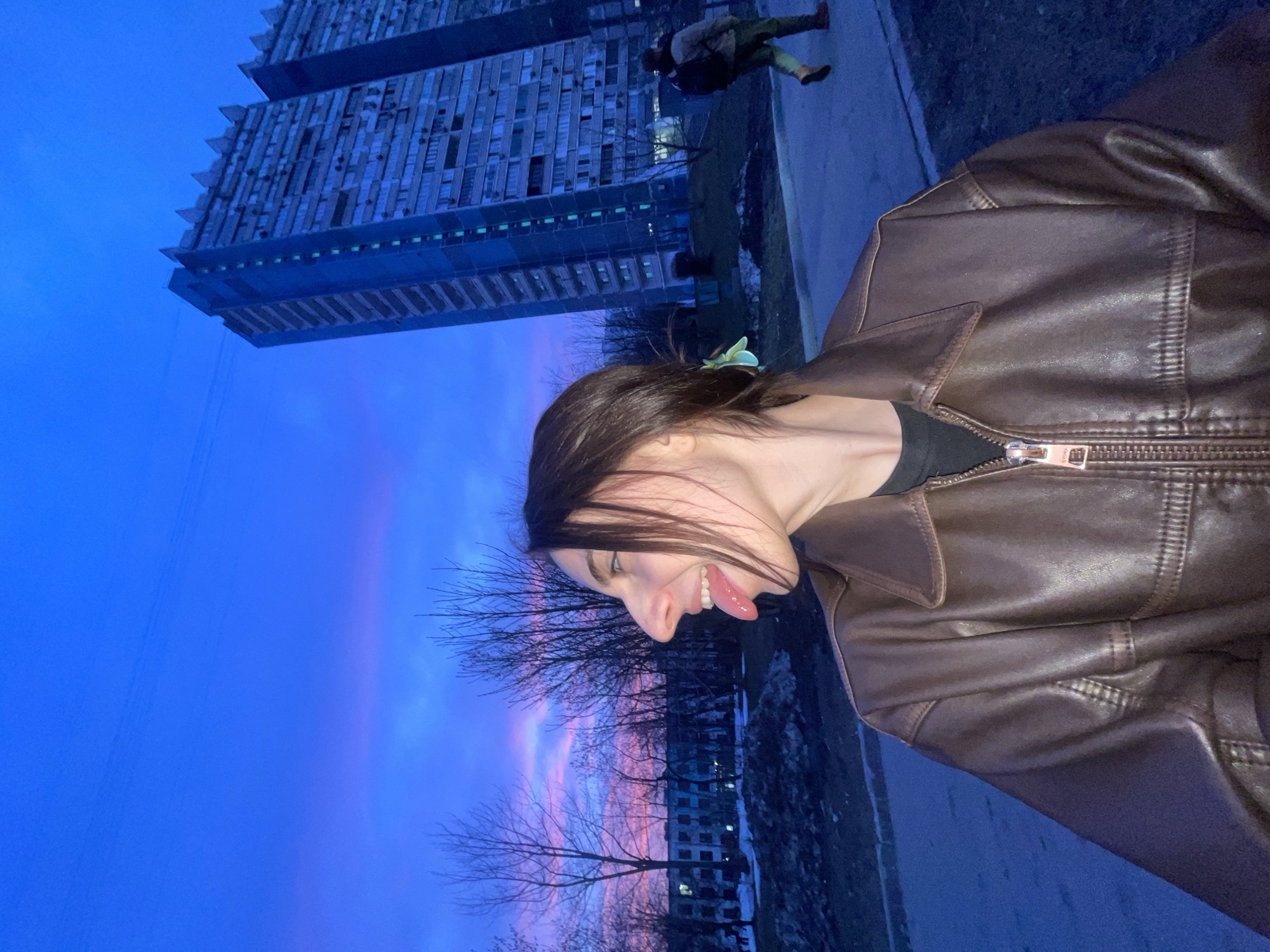 The image captures an outdoor urban scene at twilight, dominated by a vibrant blue sky transitioning to pink and purple at the horizon. In the foreground, positioned on the left, a female individual is seen in profile, looking towards the right. She has dark hair, a light green accessory in her hair, and is wearing a dark brown, likely leather, jacket over a black top. Her mouth is open, and her tongue is extended in a playful manner.
In the midground and background, on the right side of the frame, a prominent, multi-story concrete residential building stands tall, identified as one of the "House-Ships" (Дома-корабли) located at **Veshnyakovskaya Street, 22, Moscow, Russia**. This building exhibits numerous rectangular windows and balconies and features a distinctive, serrated or jagged roofline. A similar building is partially visible behind it to the right. To the right of the main subject, a male figure is observed walking away from the camera on a paved path. He wears a light-colored jacket, dark green pants, and carries a dark backpack. Bare deciduous trees are present across the midground and left background, and patches of dirty snow or ice are visible on the ground, suggesting a cold season.