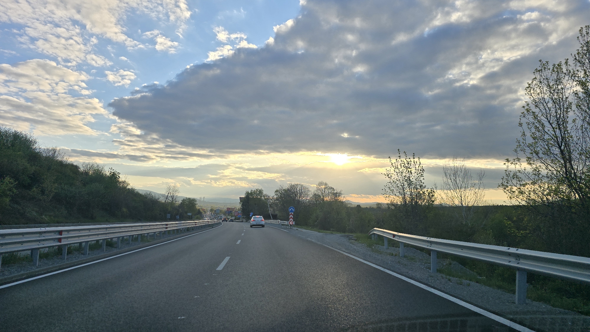 The image shows a view from a vehicle traveling on a highway near Marchaevo, Bulgaria. The road is straight, with a double white line dividing the lanes, and safety barriers on both sides. A white car is visible ahead in the same lane. The sky is filled with clouds, and the sun is partially obscured, creating a bright glow. Trees and greenery line the sides of the road. In the distance, hints of an urban area or industrial complex can be seen. The overall impression is one of a drive through a rural or semi-rural landscape during sunset or sunrise.