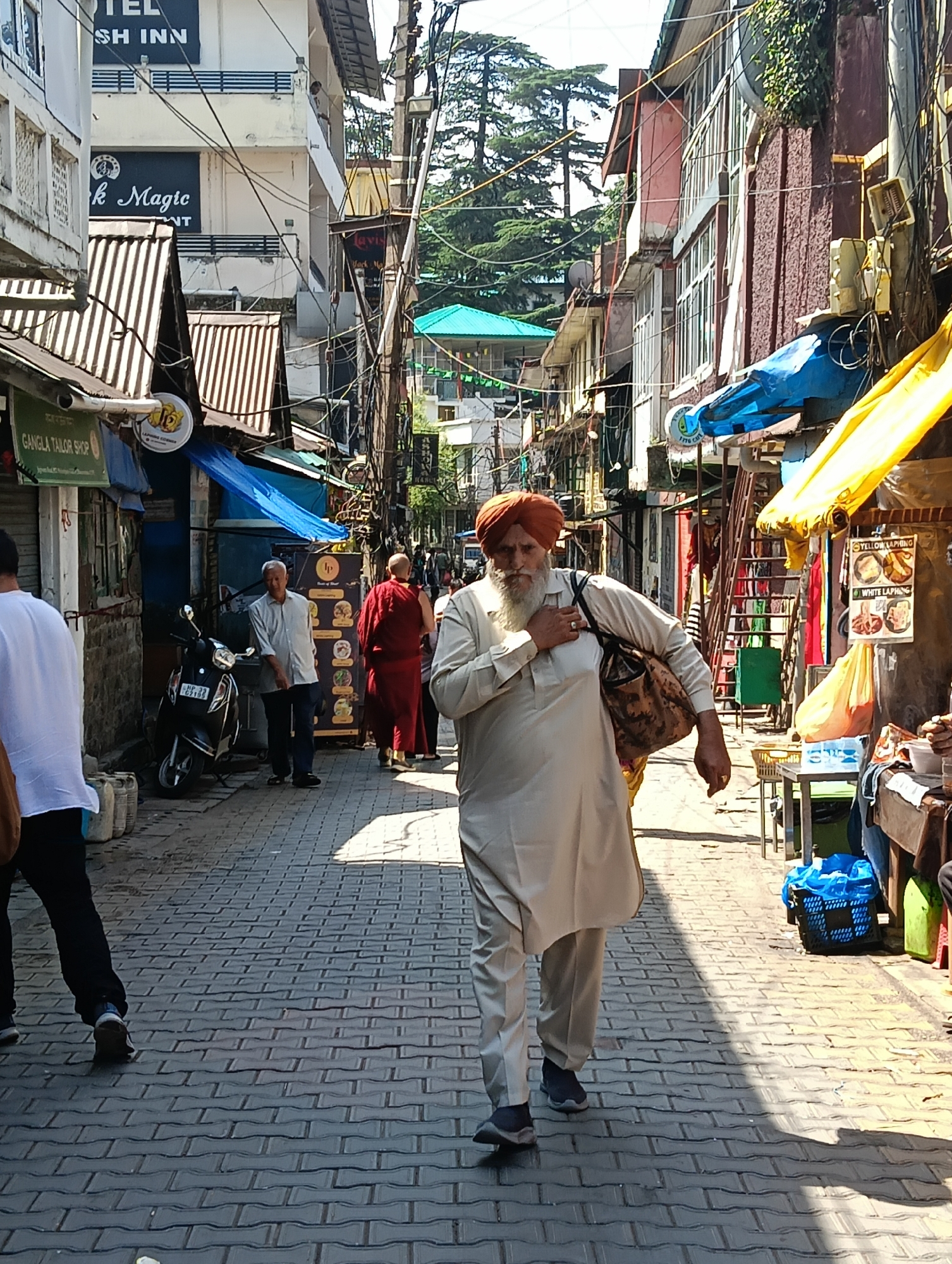 The image shows a street scene in Dharamshala, India. A Sikh man wearing a turban and traditional attire is walking towards the camera, carrying a bag over his shoulder. In the background, there are buildings with shops and signs, including "Gangla Tailor Shop". A scooter is parked on the side of the street. Other individuals are visible in the distance, and the street is paved with interlocking bricks. The environment appears to be an urban setting with a mix of commercial establishments and pedestrian activity.