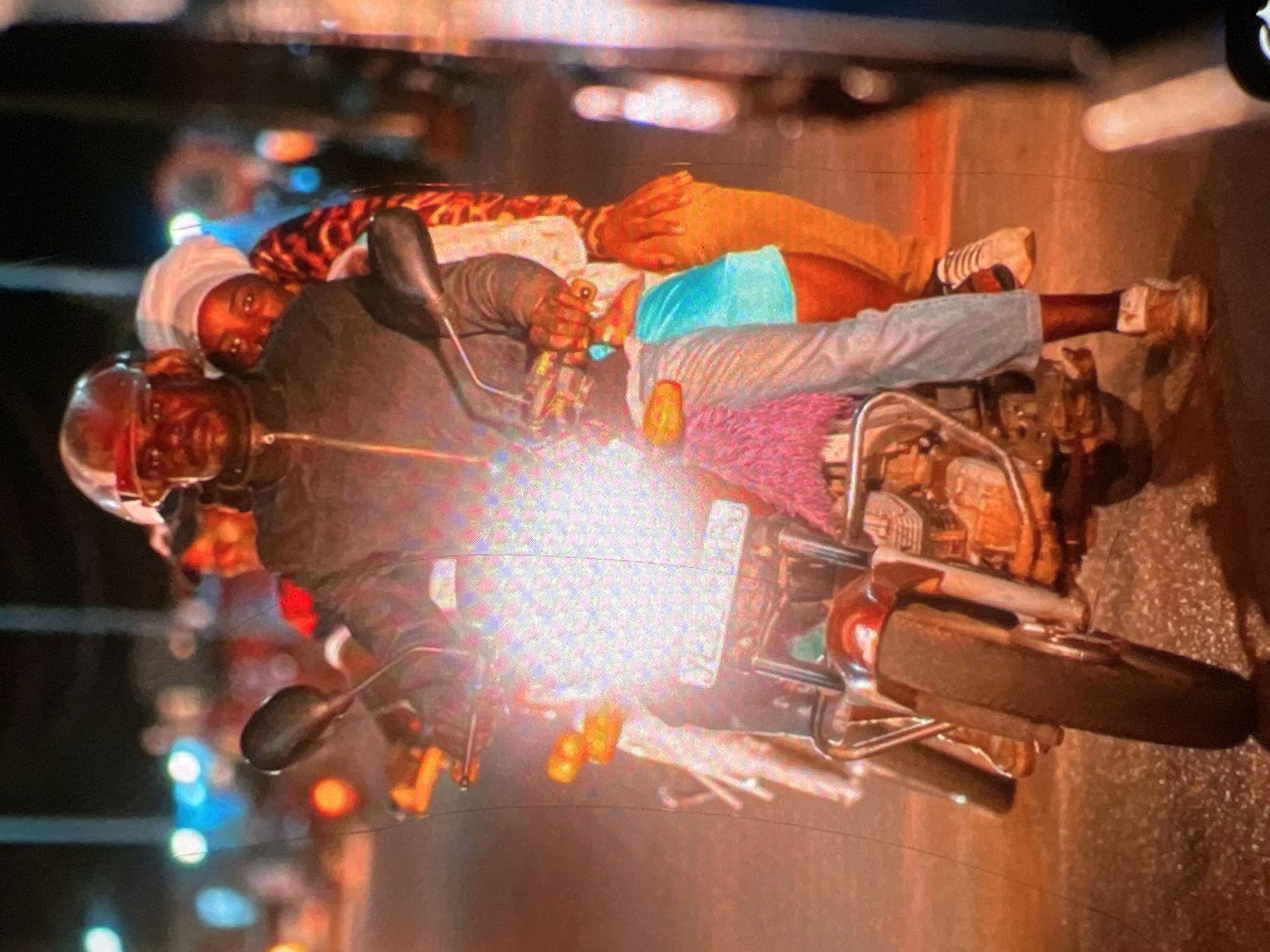 An over-the-shoulder, medium shot captures a man driving a motorcycle at night in Lagos, Nigeria, with a woman riding behind him. The driver wears a helmet, a dark jacket, and light-colored pants. The woman is dressed in a white headwrap and a vibrantly patterned outfit. The motorcycle is illuminated by its bright headlights. In the background, blurred lights suggest a busy urban environment.