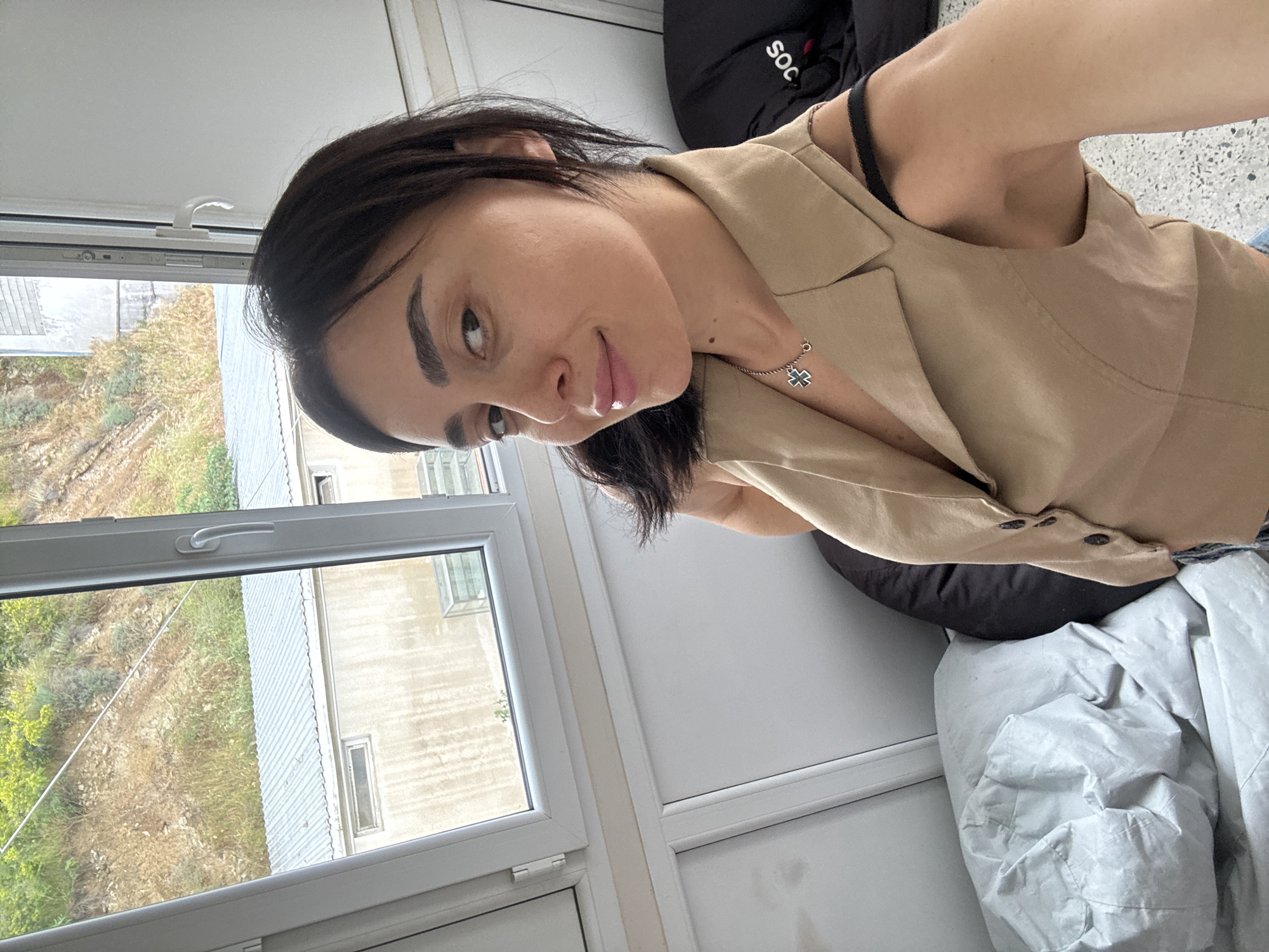The image shows a young woman with dark hair and light skin posing for a selfie indoors. She is wearing a tan vest over a black bra and a necklace with a cross pendant. She is looking towards the camera with a slight smile. In the background, there is a window with a view of a hillside covered in dry grass and some buildings. The room appears to be a residential or office space, with furniture and belongings visible. Based on the architectural style and the landscape, the location is consistent with the city of Tbilisi, Georgia. The image quality is good, and the lighting is natural.