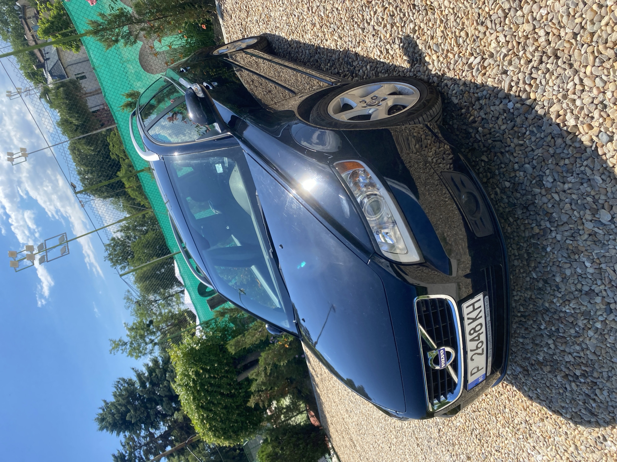 A black Volvo V50 station wagon is parked on a gravel lot. The car is facing forward and slightly to the left. It has a Bulgarian license plate that reads "P 2648KH". Behind the car, there is a green tennis court fence and some trees. The sky is blue with some clouds. The scene is likely in Varna, Bulgaria, as indicated by the license plate and the style of the buildings in the background. The car is in good condition, with no visible damage. The lighting suggests it is daytime and sunny.