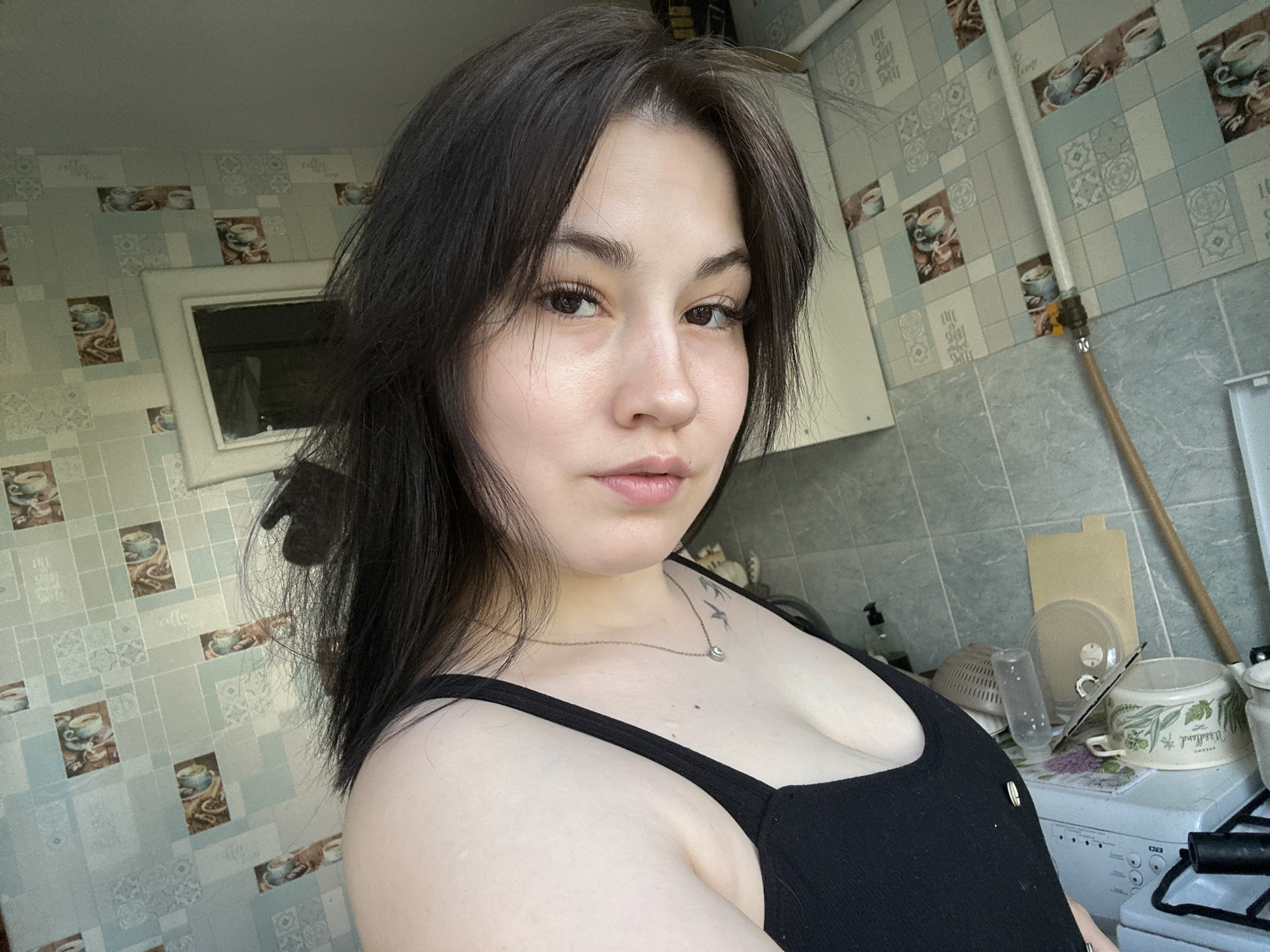 A medium close-up shot captures a woman with dark hair and fair skin, wearing a black tank top and a silver necklace. She appears to be indoors, possibly in a kitchen setting. The background includes patterned wallpaper with images of coffee cups and kitchen tiles, a window with a white frame, and various kitchen items such as cookware and utensils on a stove. It is not possible to confirm the location to be the city of Chaikovsky, Russia, without additional information.